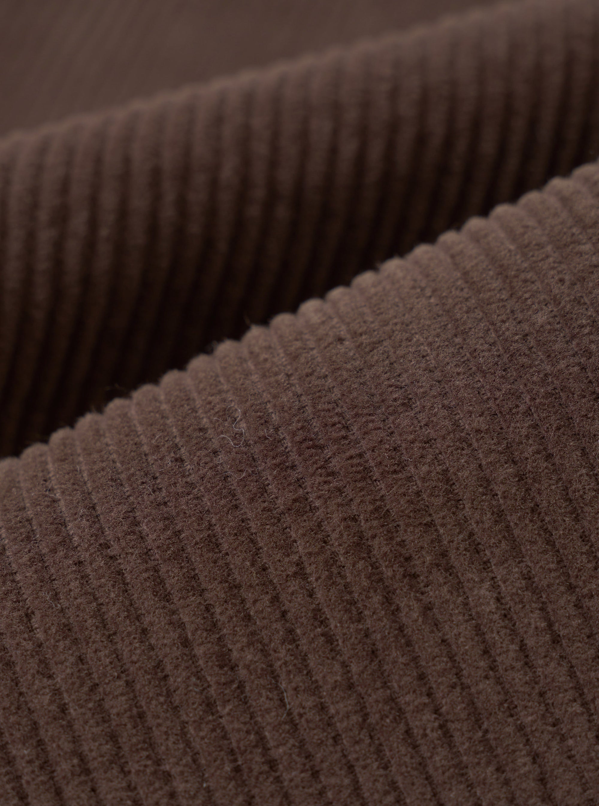 Close-up detail shot of cacao brown corduroy fabric, showing wide, clearly defined wales running diagonally across the frame. The raised ridges and textured valleys of the cord are sharply in focus, highlighting the soft, dense pile and matte finish of the material.
