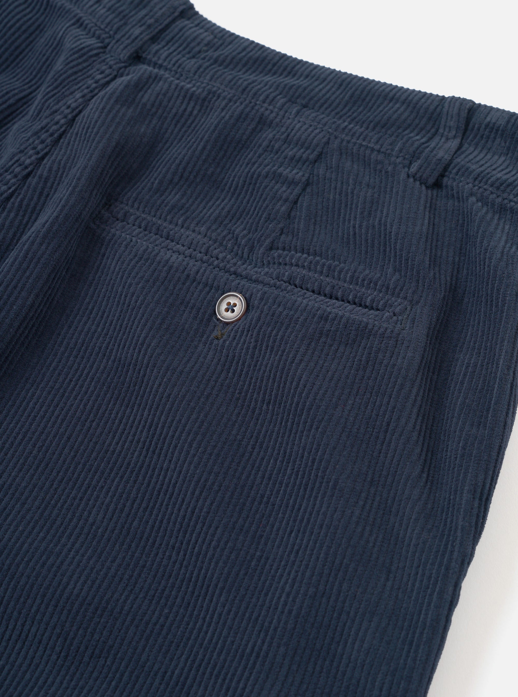 Close-up detail of the rear of dark navy corduroy military chinos, shot flat on a white studio surface. A welt back pocket with a single four-hole button fastening is visible. The wide-wale corduroy texture runs vertically across the fabric. The waistband sits at the top of the frame.