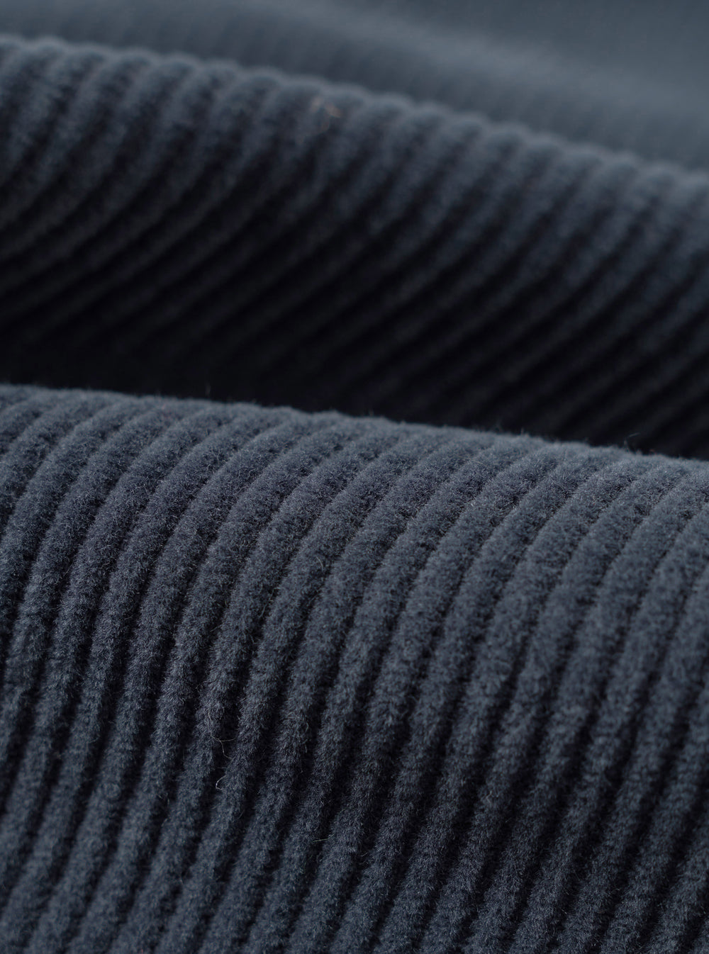 Close-up fabric detail of the dark navy cord military chino, showing wide-wale corduroy texture with raised ridges running diagonally across the frame. Two overlapping layers of the fabric are visible, highlighting the deep navy tone and soft, dense pile of the cord.