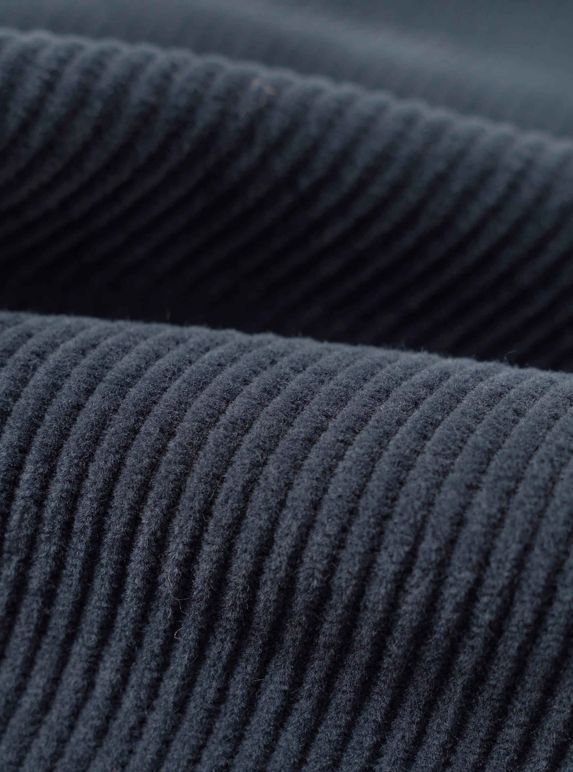 Close-up fabric detail of the dark navy cord military chino, showing wide-wale corduroy texture with raised ridges running diagonally across the frame. Two overlapping layers of the fabric are visible, highlighting the deep navy tone and soft, dense pile of the cord.