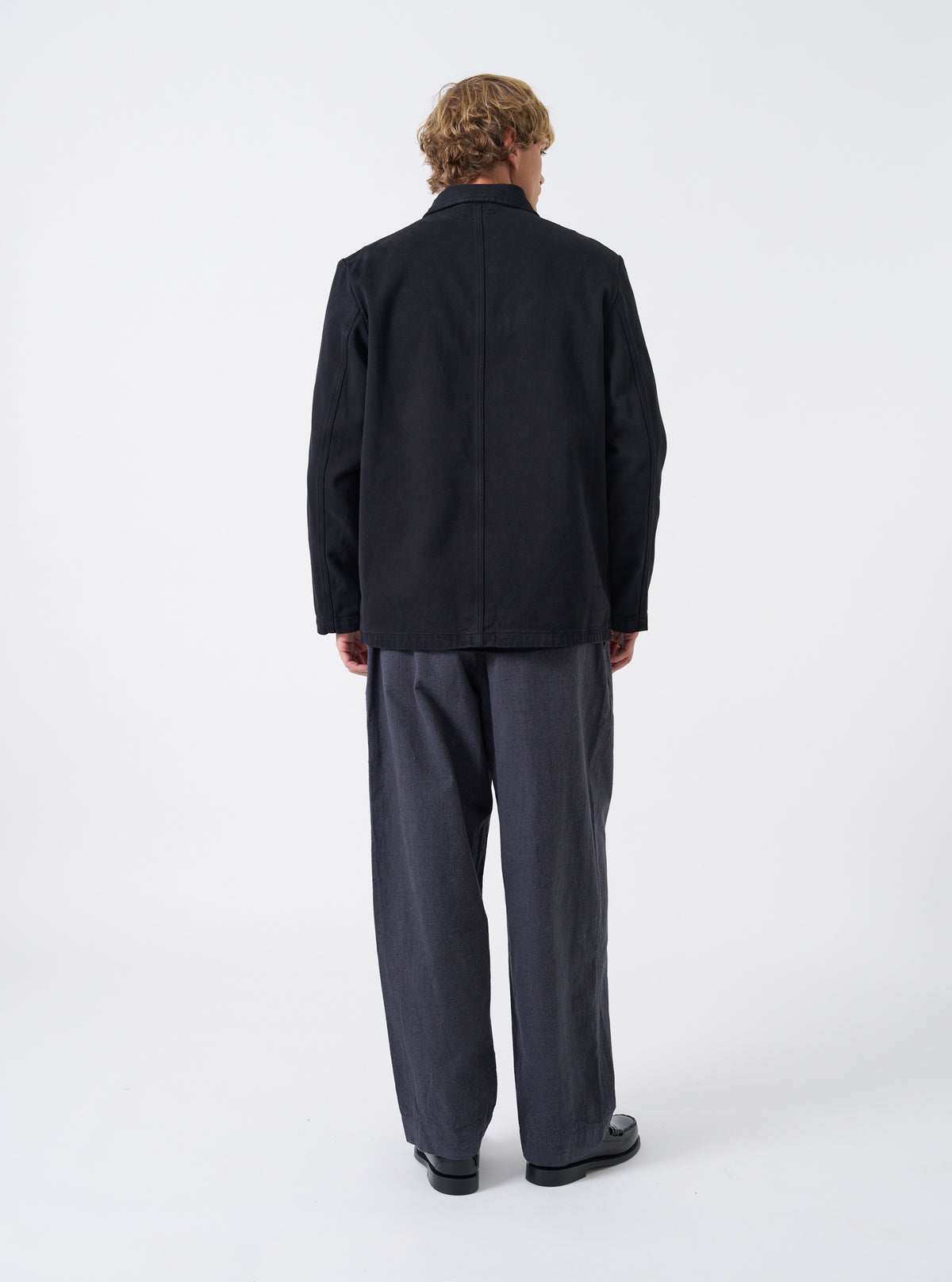 A male model wears the Black Dexter Twill OG Bakers Chore Jacket unbuttoned over a shirt, standing against a neutral studio background. The view details the open front layer, collar, and comfortable overall silhouette.