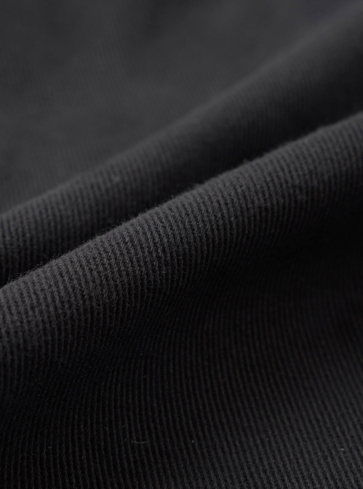 A close-up fabric swatch view displaying the rich texture and fine weave of the organic cotton twill fabric from the Black Dexter Twill Grad Chino. The frame is filled entirely with dark black twill fabric detail.