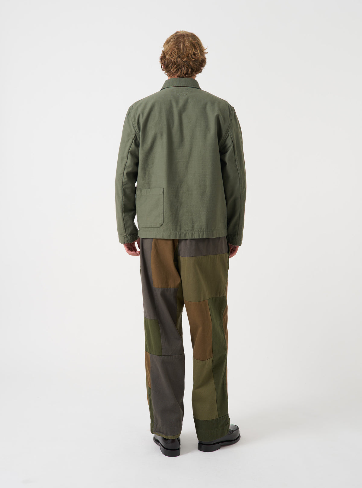 A male model wears the Olive Slub Sateen Ives Jacket, captured in a three-quarter side pose against a plain light grey studio backdrop, highlighting the sleeve length, boxy cut, and side profile view.