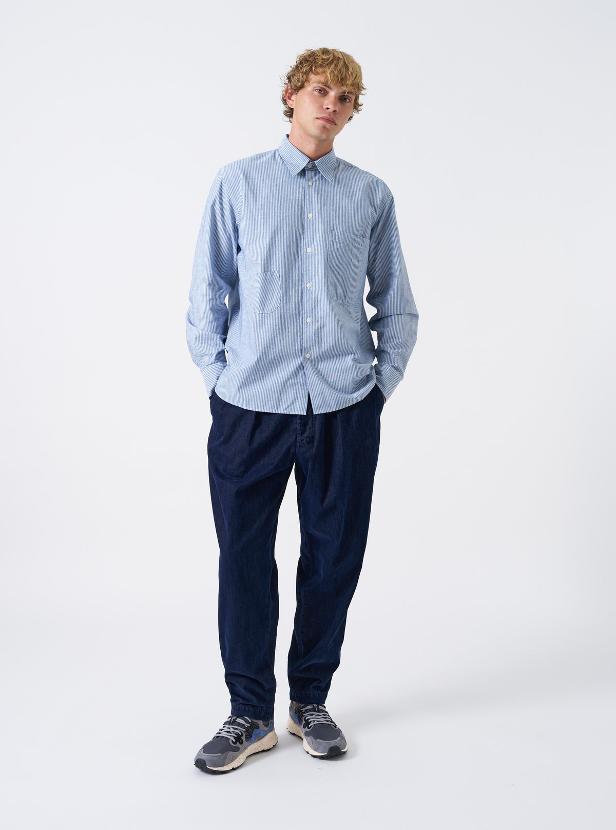 A male model wears the Blue Montgomery Stripe Station Shirt buttoned up, posing against a neutral studio backdrop. The full-length shot illustrates the blue vertical stripe pattern, classic collar, chest pockets, and comfortable relaxed fit.