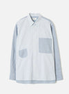 Mix Stripes WM Station Shirt in blue and white pinstripe cotton, flat on a light grey studio backdrop. Point collar with a brand label at the neckline, buttoned placket with cream buttons, two oversized patch pockets in contrasting stripe panels, long sleeves with single-button cuffs, and a curved hem.