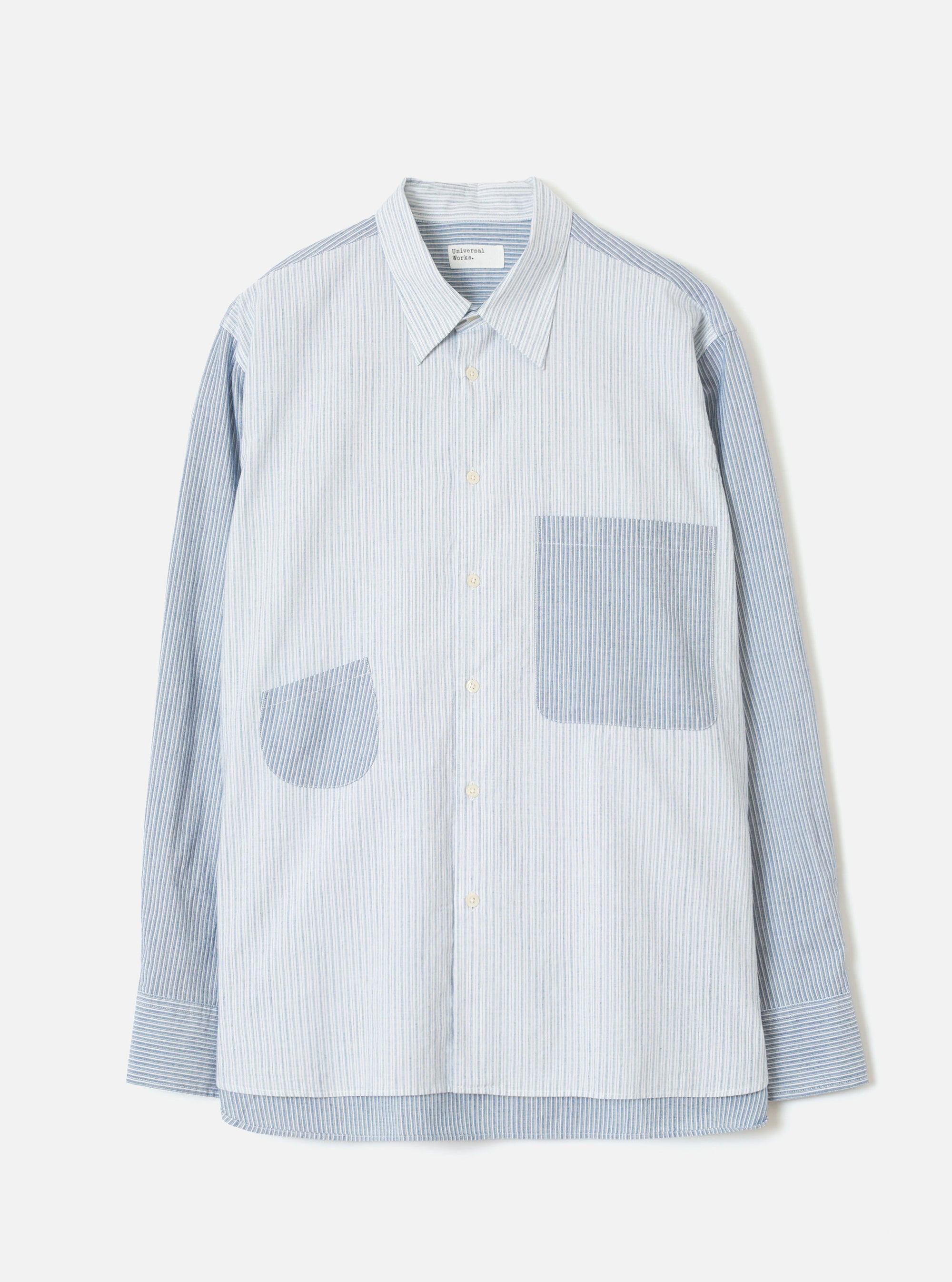 Mix Stripes WM Station Shirt in blue and white pinstripe cotton, flat on a light grey studio backdrop. Point collar with a brand label at the neckline, buttoned placket with cream buttons, two oversized patch pockets in contrasting stripe panels, long sleeves with single-button cuffs, and a curved hem.