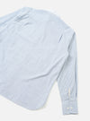 Flat lay of a light blue and white mix-stripe station shirt on a white studio surface. The shot crops the upper-left corner, showing the back panel, one long sleeve, and a rounded hem. The cuff is buttoned with two small white buttons and features a barrel-style finish. A small woven label is visible at the hem seam.