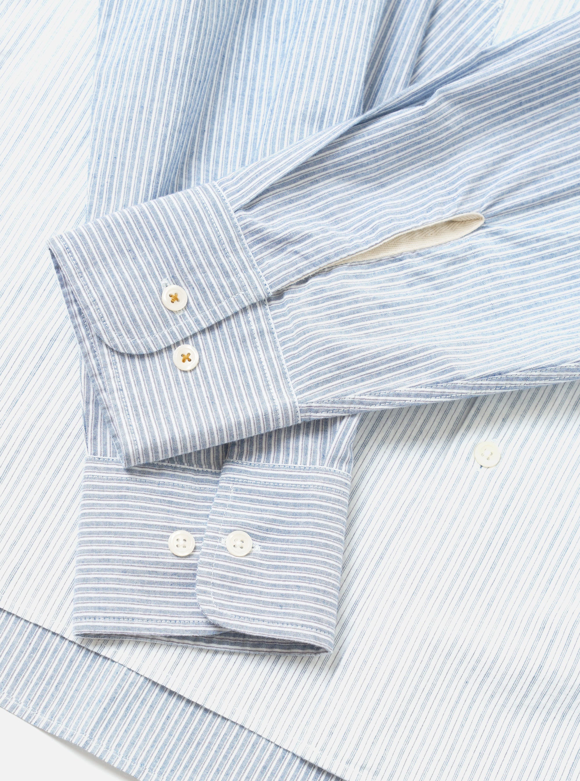 Close-up flat lay of the Mix Stripes WM Station Shirt in blue and white. Two barrel cuffs are shown overlapping, each fastened with two mother-of-pearl buttons. The woven fabric features fine vertical blue and white stripes with a slightly textured surface. Shot on a white studio surface.