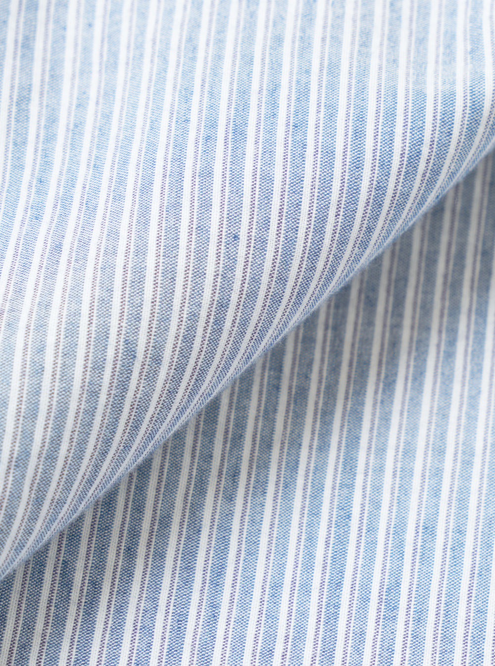 Close-up fabric detail of the Mix Stripes Station Shirt, showing a woven blue and white striped textile with a fine pin-stripe pattern. The fabric has a chambray-like texture with visible slub threads. A diagonal fold runs across the frame, revealing the fabric's drape and weight.