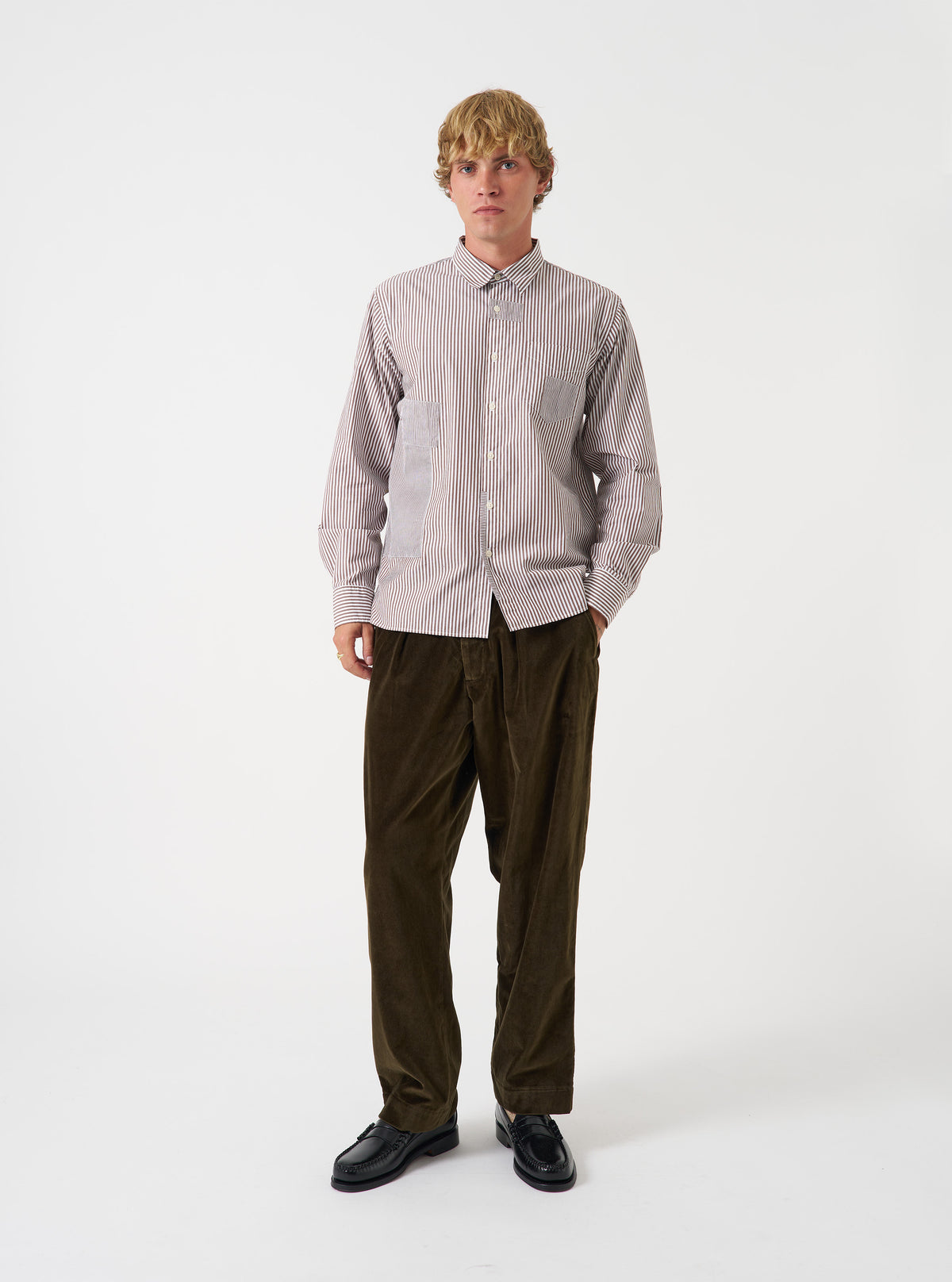 A male model wears the Brown Queens Cotton Patched Shirt standing against a neutral studio backdrop. The front view highlights the button-down collar, chest pocket, and patchwork panel details across the garment.