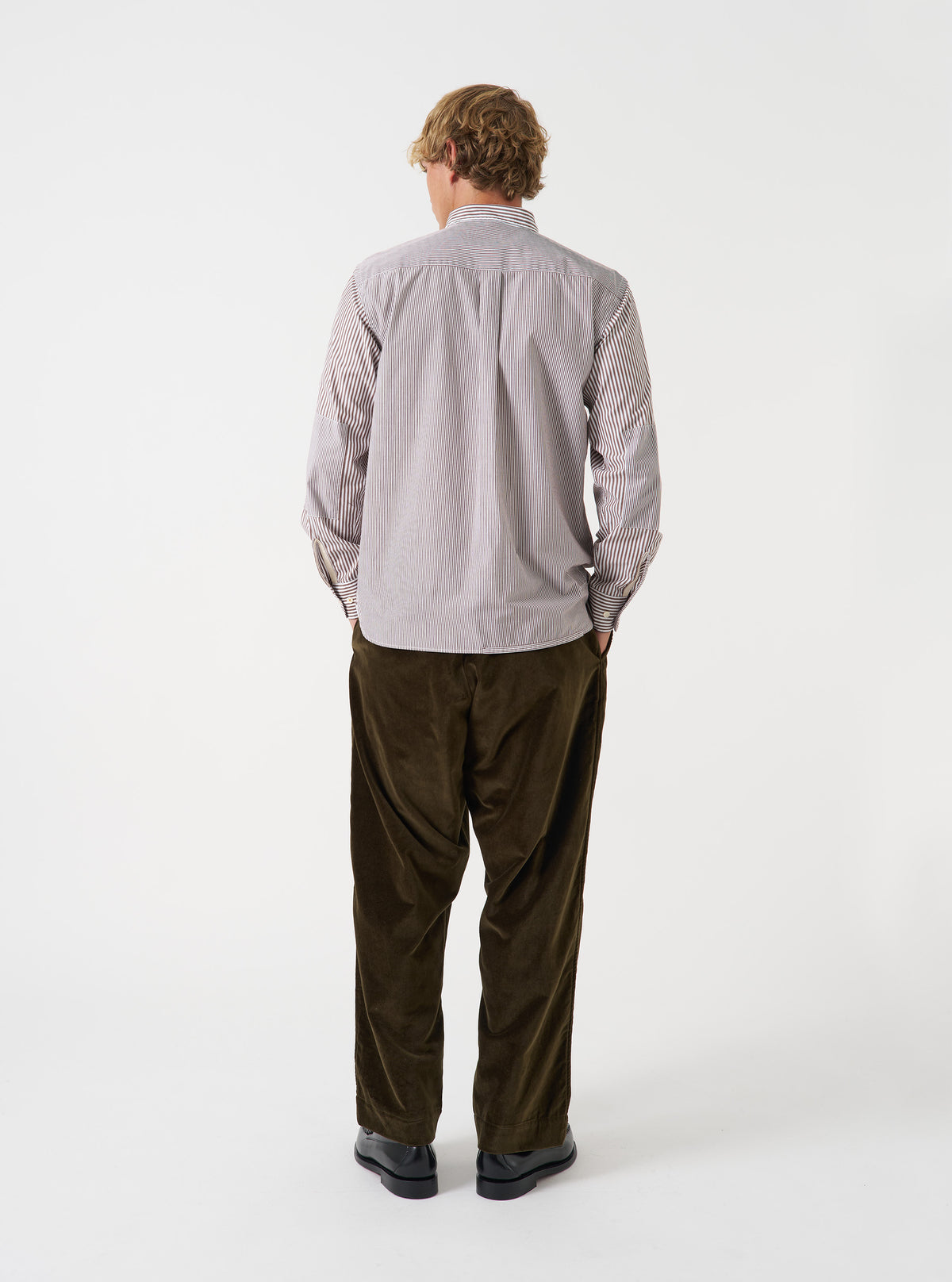 A male model wears the Brown Queens Cotton Patched Shirt posed against a plain studio background. This shot showcases the relaxed fit, woven stripe texture, and distinct patched design from a side angle.