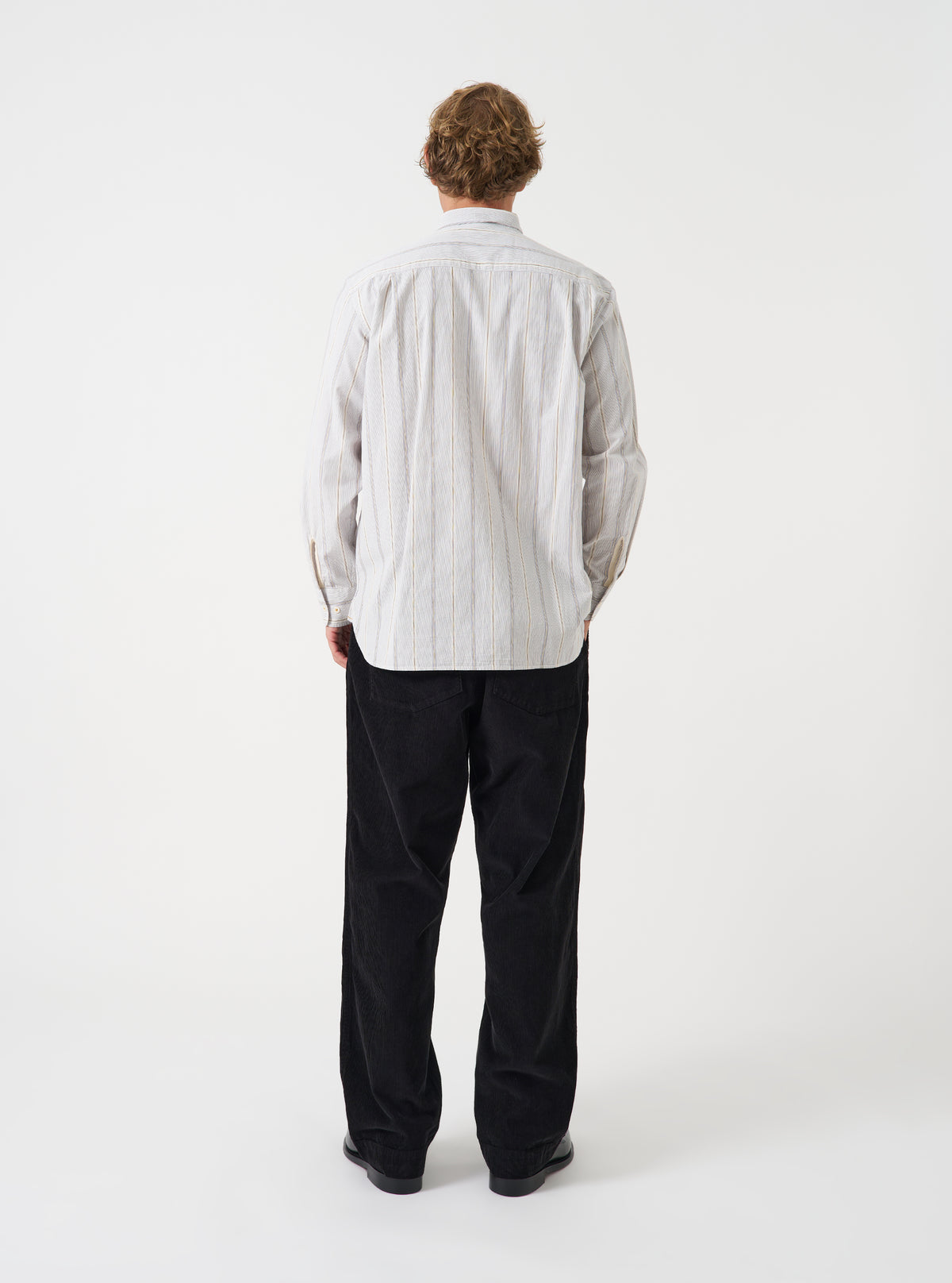 A male model wears the Dove Monk Organic Stripe Square Pocket Shirt in a back view shot against a neutral studio backdrop. The photo details the subtle striped fabric and clean rear silhouette of the shirt.