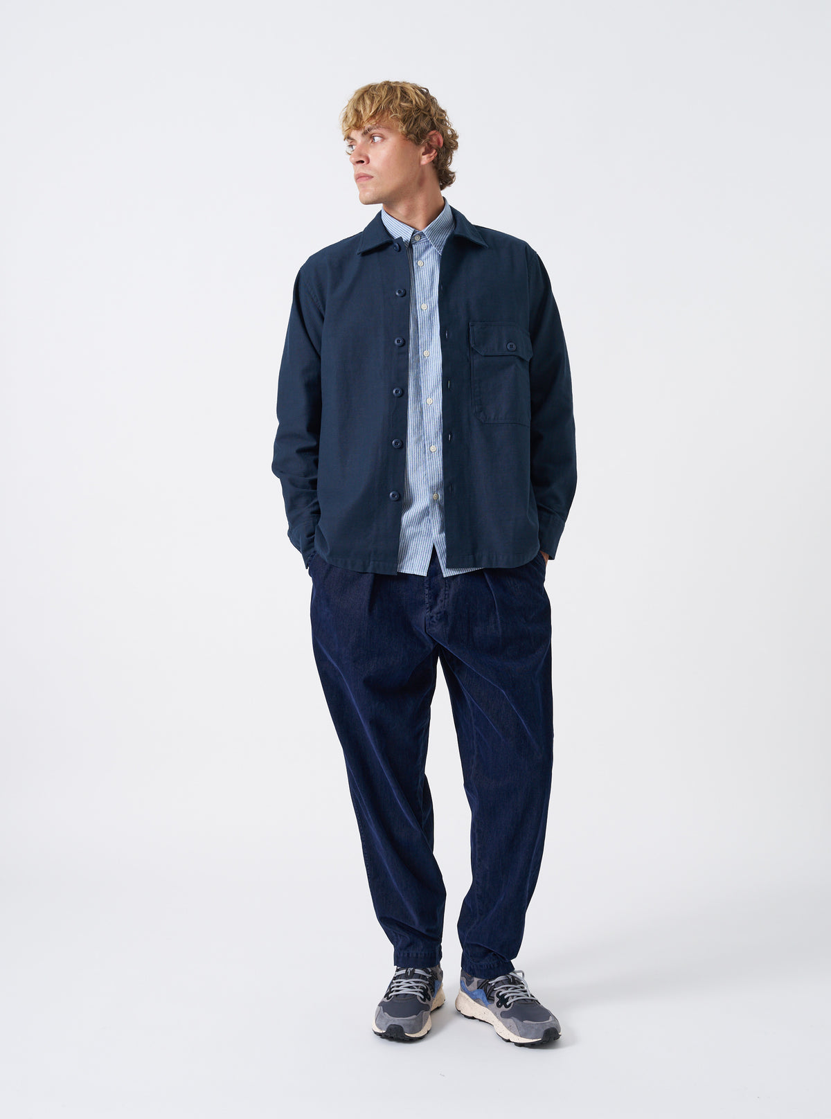 A male model wears the Navy Slub Sateen Fatigue Shirt unbuttoned over a top, posing against a neutral studio backdrop. The front view demonstrates the relaxed drape, chest pockets, and overall fit.