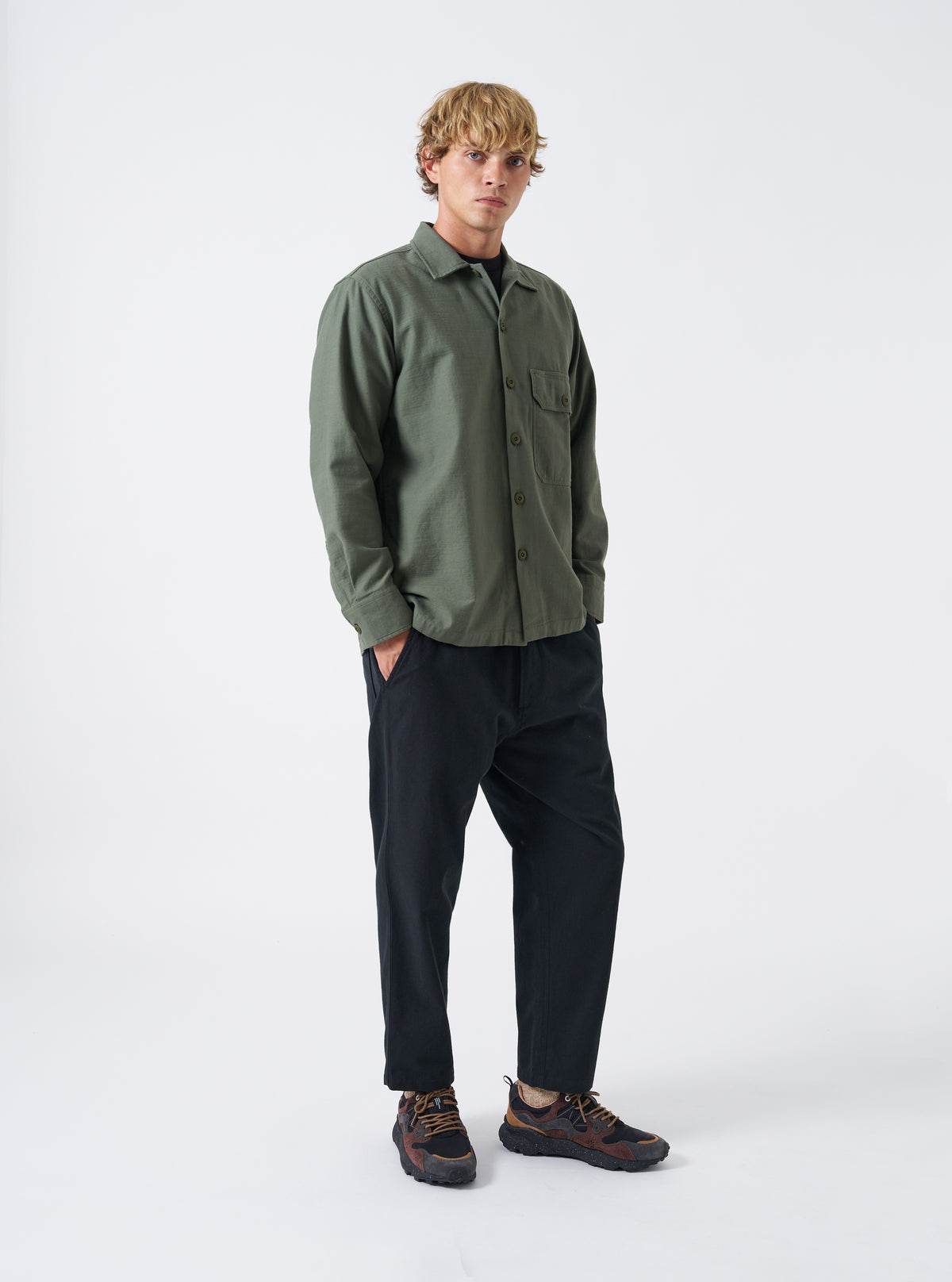 A male model wears the Olive Slub Sateen Fatigue Shirt in a front view shot against a neutral studio background. The photo showcases the olive green color, casual layering style, and chest pockets.