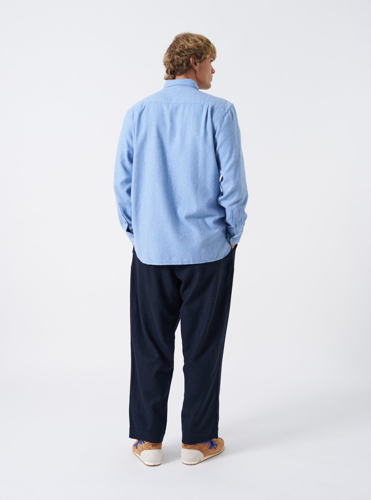 A male model poses in a studio setting wearing the Sky Blue Monty Cotton Tencel Standard Shirt unbuttoned over a plain top. The side-angled shot highlights the casual drape, chest pocket detail, and sleeve cuffs.