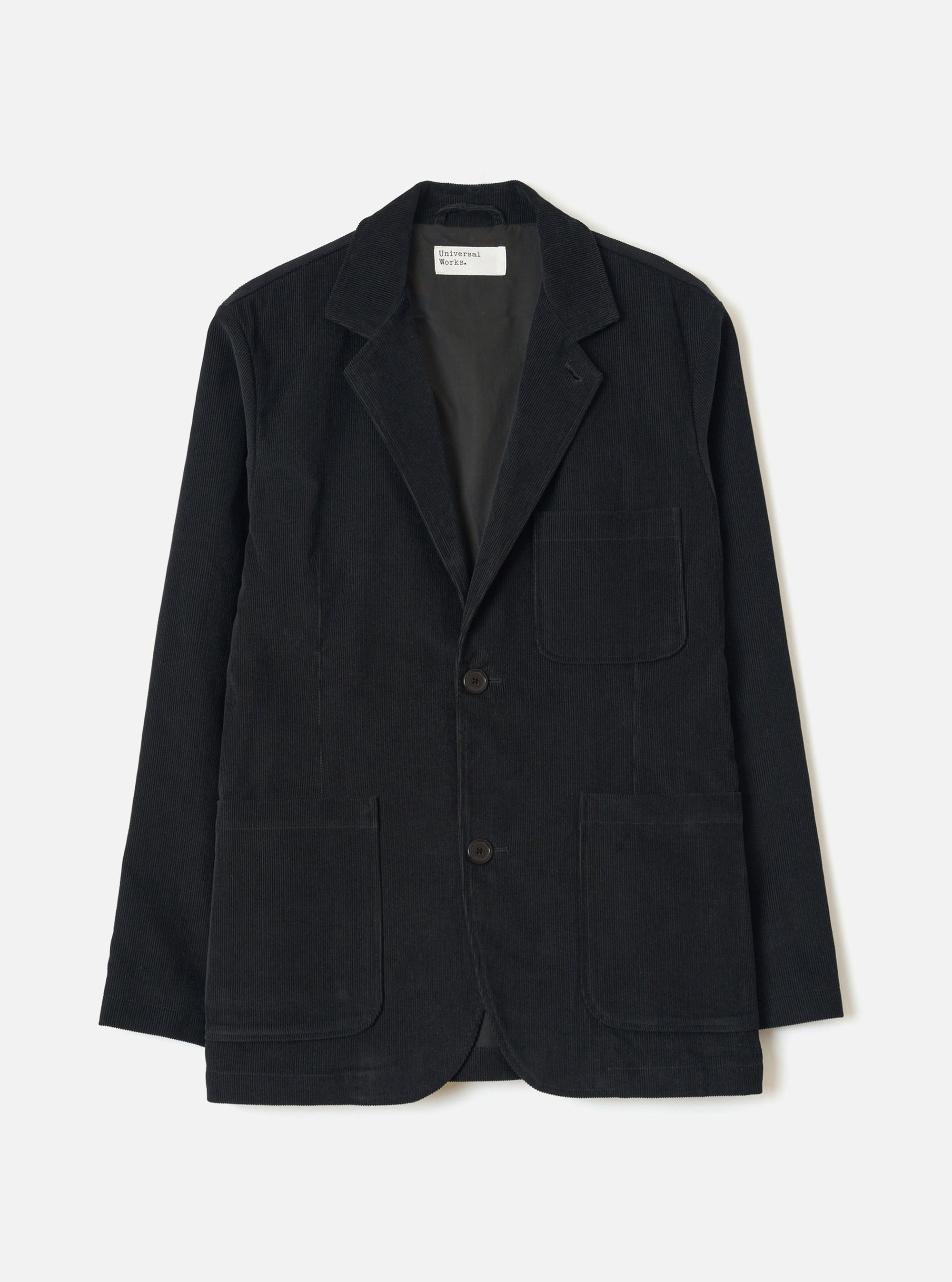 Black corduroy two-button jacket laid flat on a light grey studio backdrop. Notched lapels, open front, two large patch pockets at the hip, one chest patch pocket, dark horn-effect buttons, and full-length sleeves. A Universal Works woven label is visible at the collar. Fine cord texture throughout.