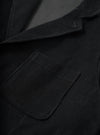 Close-up detail of the black corduroy two button jacket, showing the notched lapel and a rounded patch pocket with topstitching. The wide-wale cord texture is clearly visible across the body and lapel. A dark lining is glimpsed at the open collar. No model or mannequin present.