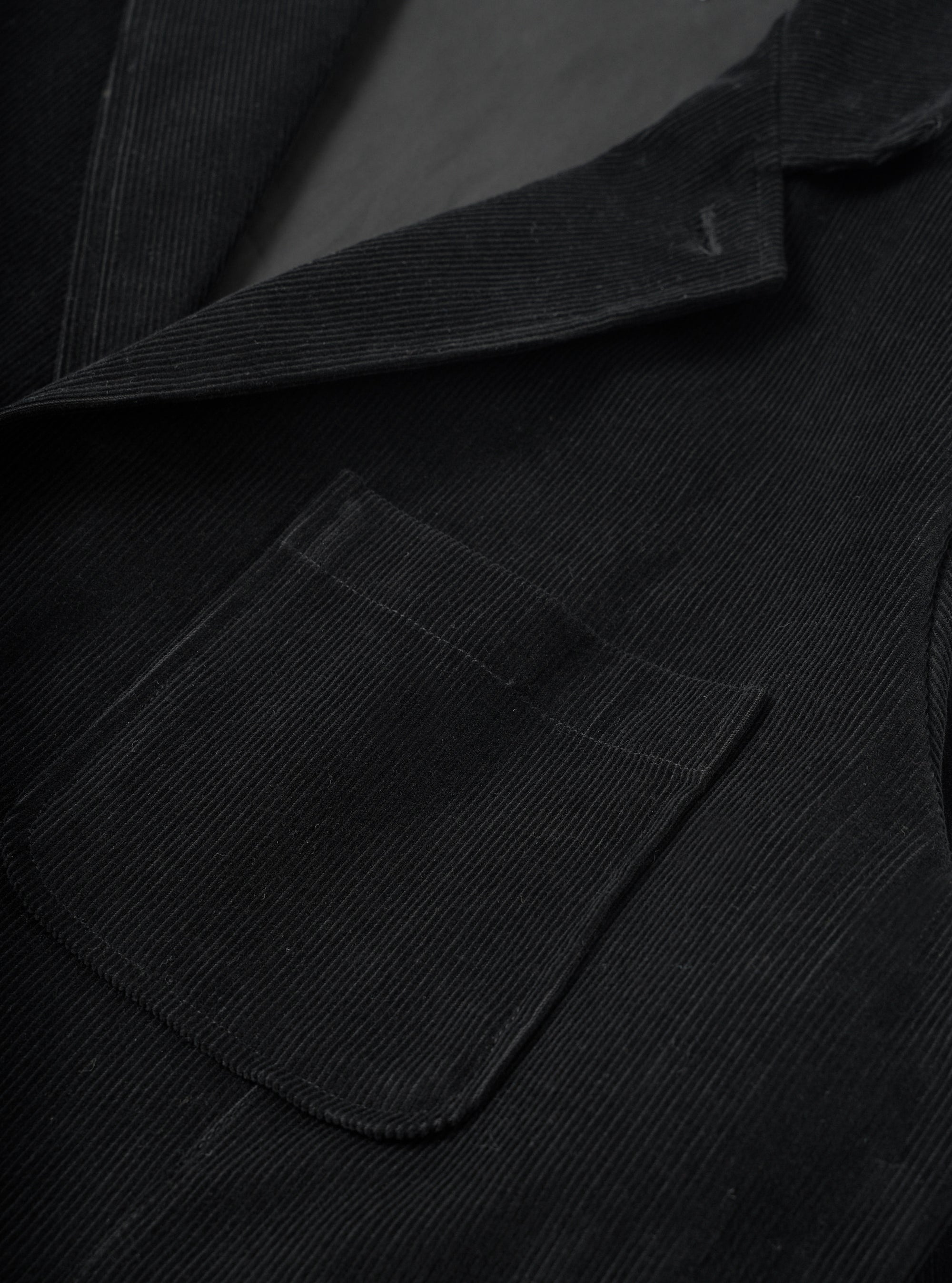 Close-up detail of the black corduroy two button jacket, showing the notched lapel and a rounded patch pocket with topstitching. The wide-wale cord texture is clearly visible across the body and lapel. A dark lining is glimpsed at the open collar. No model or mannequin present.