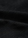 Close-up detail shot of black corduroy fabric showing wide, evenly spaced wales with a soft, textured pile. The fabric is folded to highlight the depth and structure of the cord. Dark studio background. No garment construction details visible.