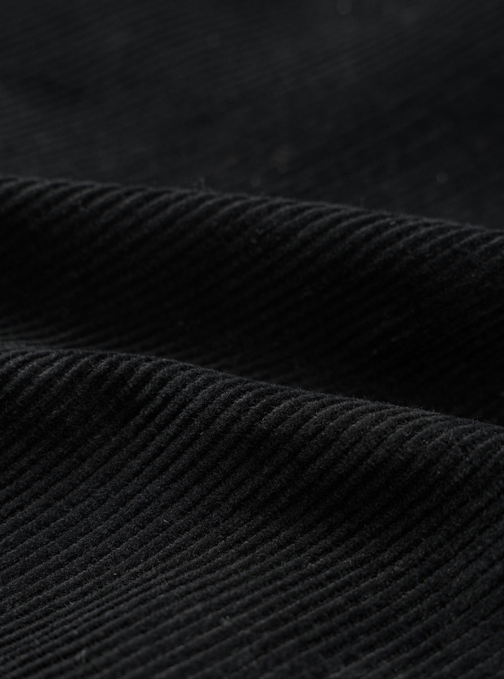 Close-up detail shot of black corduroy fabric showing wide, evenly spaced wales with a soft, textured pile. The fabric is folded to highlight the depth and structure of the cord. Dark studio background. No garment construction details visible.