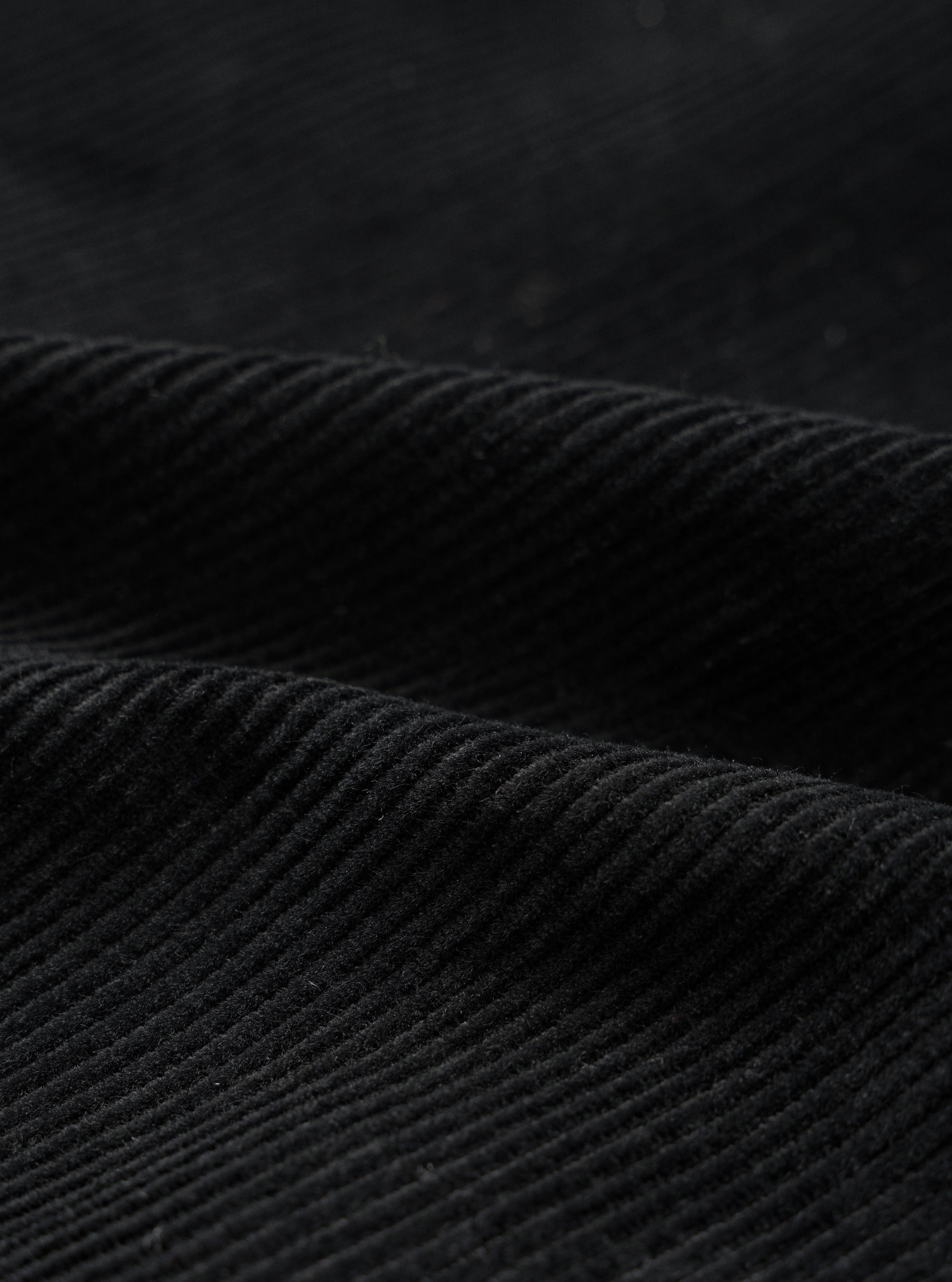 Close-up detail shot of black corduroy fabric showing wide, evenly spaced wales with a soft, textured pile. The fabric is folded to highlight the depth and structure of the cord. Dark studio background. No garment construction details visible.