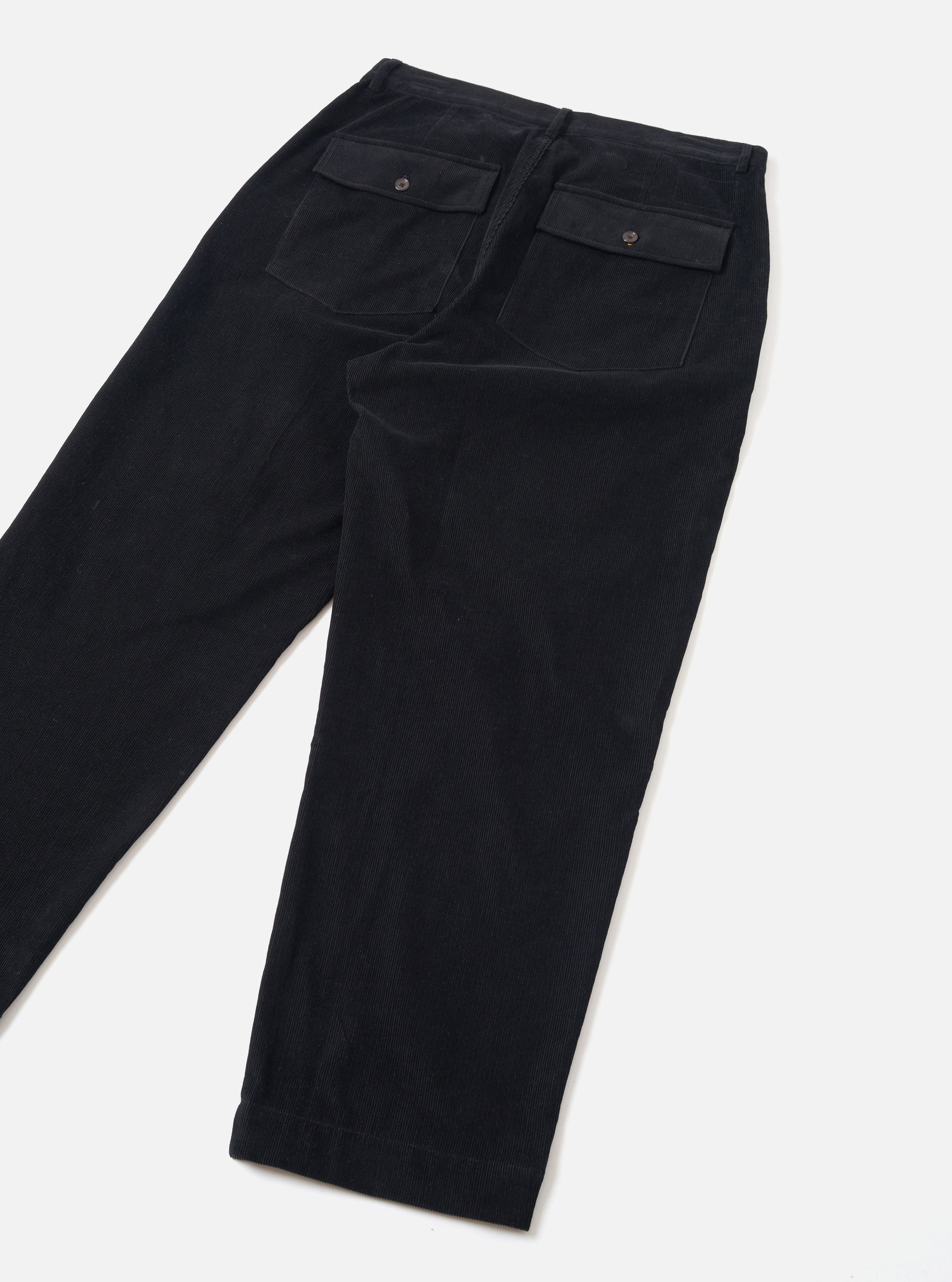 Black corduroy grad chino trousers laid flat on a light grey surface, showing the back. Fine-wale cord texture throughout. Two large patch pockets with flap closures and single metal press-stud buttons sit at the waistband. Tapered leg with full-length hem visible at the cuff.
