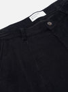 Close-up flat lay of Universal Works black corduroy Grad Chino trousers on a light grey backdrop. Wide-wale cord texture is clearly visible. The waistband, a single belt loop, and a gunmetal button fastening are shown. A white woven label reads "Universal Works." is visible at the top.