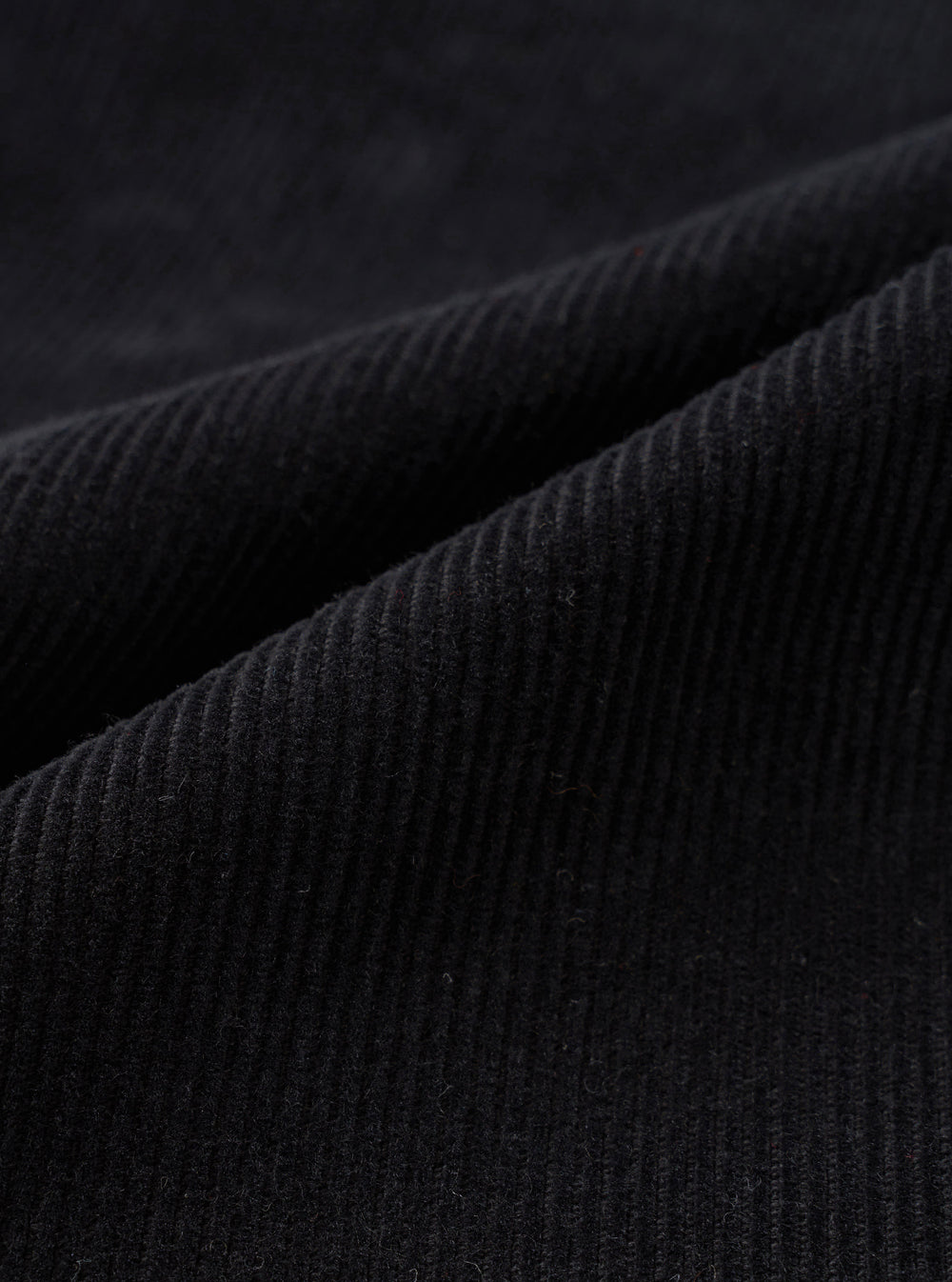 Close-up detail shot of black corduroy fabric showing wide-wale cord texture. The parallel ridges and soft pile of the material are clearly visible across the draped folds of the Black Cat Cord Grad Chino. No background is visible.