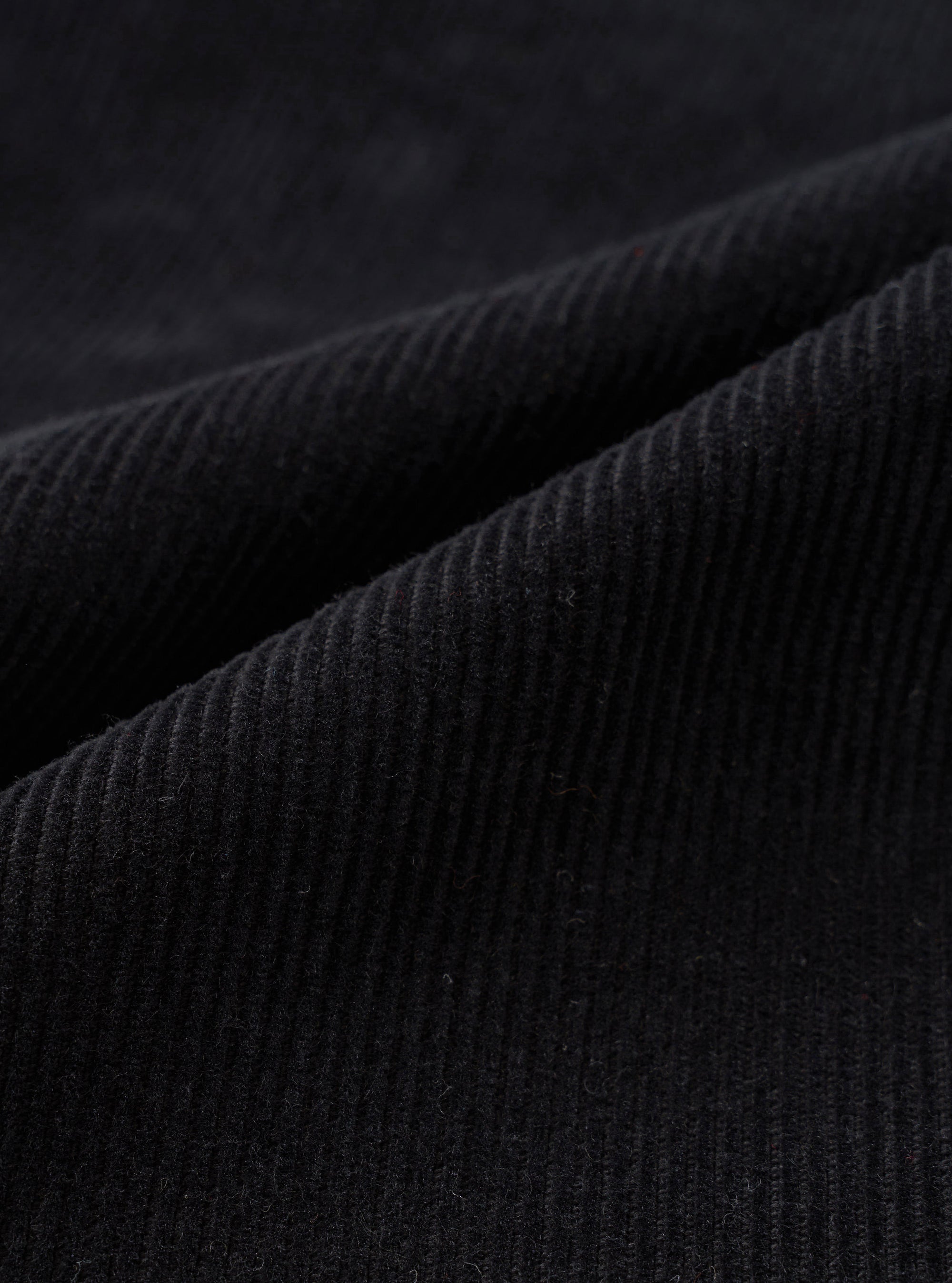 Close-up detail shot of black corduroy fabric showing wide-wale cord texture. The parallel ridges and soft pile of the material are clearly visible across the draped folds of the Black Cat Cord Grad Chino. No background is visible.