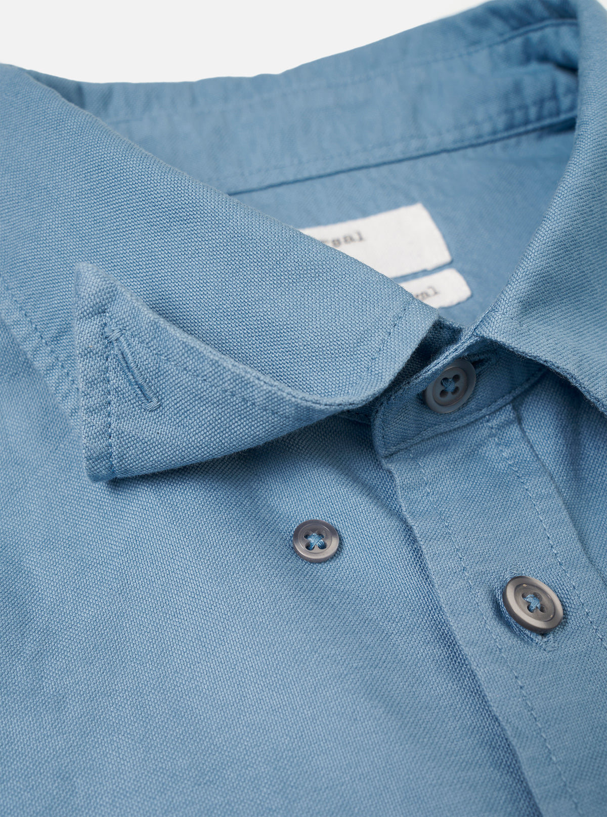 A close-up shot of the Mid Blue R.Oxford Lazy Day Shirt showing the button-down collar, front placket buttons, and asymmetric chest pocket details set against a plain off-white background.