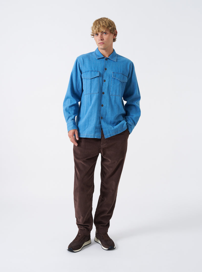 A male model wears the Washed Indigo Recycled Herringbone Attendant Shirt buttoned up over a plain t-shirt, standing against a neutral studio background. The full shot highlights the utility fit and overall styling.