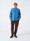 A male model wears the Washed Indigo Recycled Herringbone Attendant Shirt buttoned up over a plain t-shirt, standing against a neutral studio background. The full shot highlights the utility fit and overall styling.