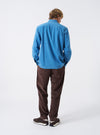 A male model wears the Washed Indigo Recycled Herringbone Attendant Shirt unbuttoned in a three-quarter pose against a plain studio background. The image showcases the shirt's relaxed layer fit and soft collar structure.