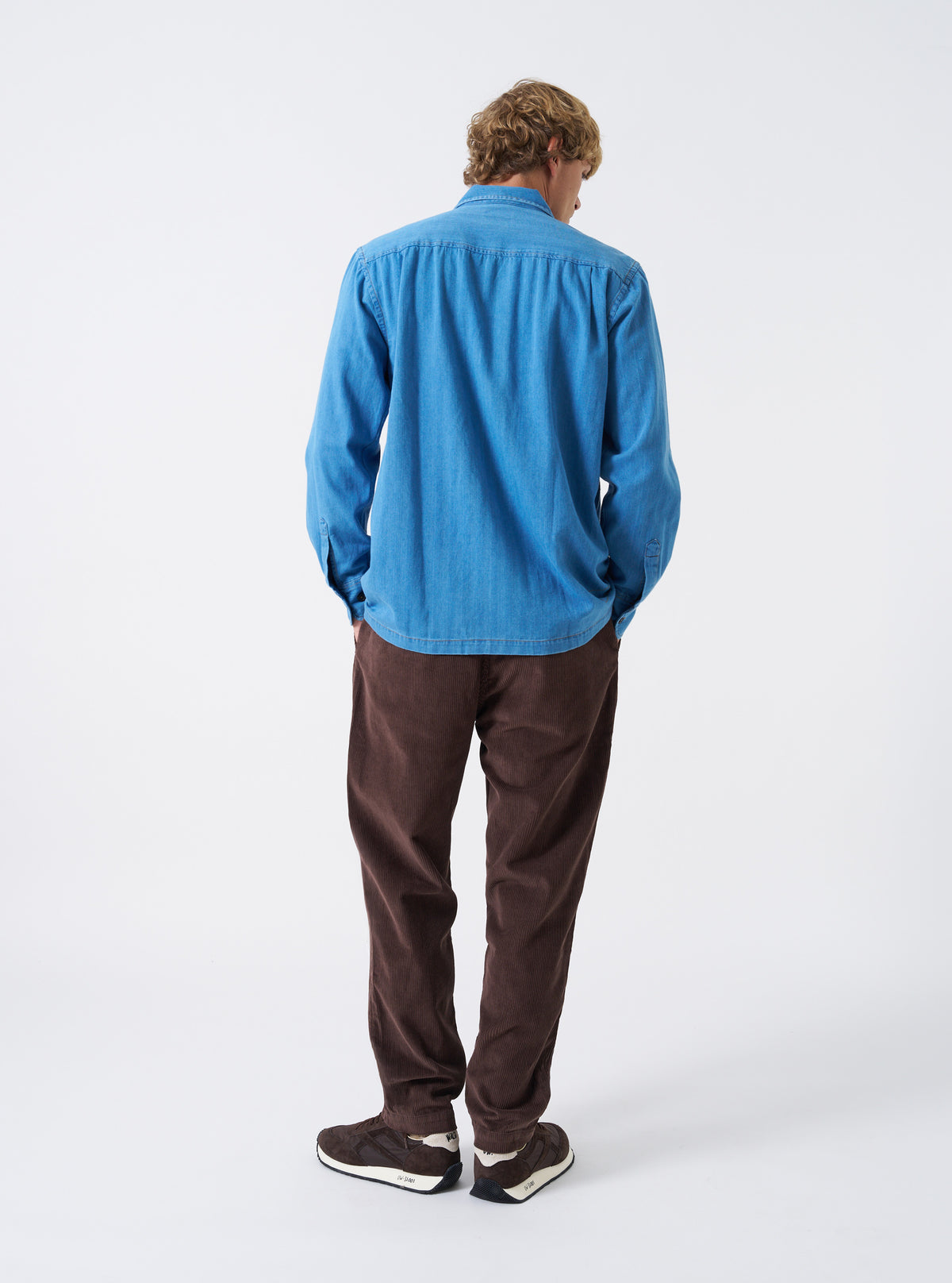 A male model wears the Washed Indigo Recycled Herringbone Attendant Shirt unbuttoned in a three-quarter pose against a plain studio background. The image showcases the shirt's relaxed layer fit and soft collar structure.