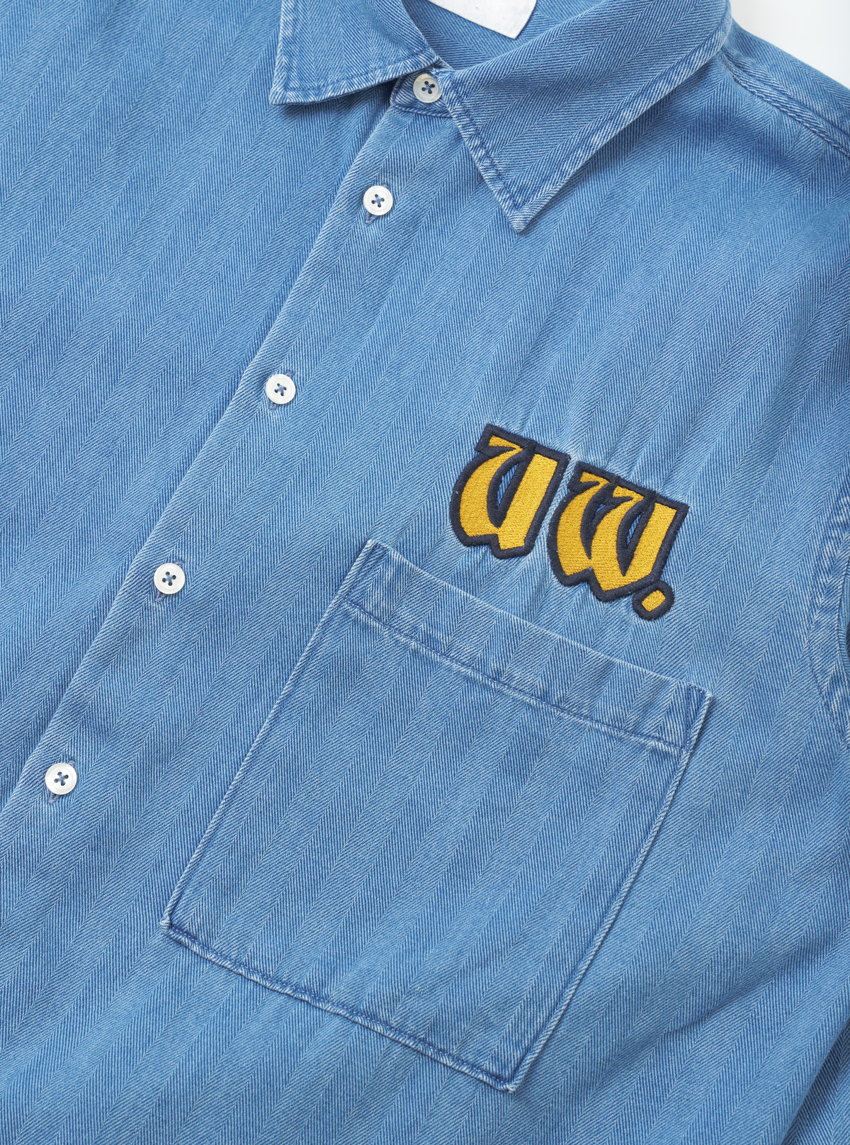 Close-up detail shot of the Washed Indigo Recycled Herringbone Patch Lucky Shirt on a plain background. The view focuses on the woven recycled cotton fabric, clean stitching, and classic button placket detailing.