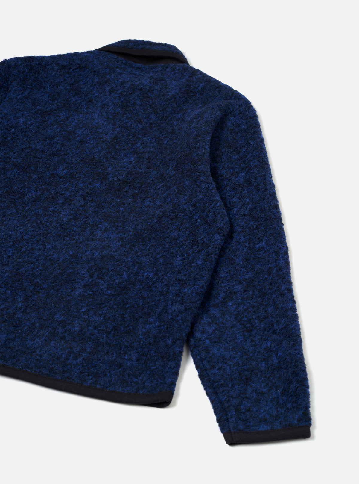 Close-up rear view of the blue boucle style fleece Lancaster jacket on a plain light grey studio backdrop. The textured boucle style fabric shows a navy and cobalt mottled finish. A dark navy collar, cuff trim, and hem band are visible, contrasting with the main body. One sleeve is shown in full.