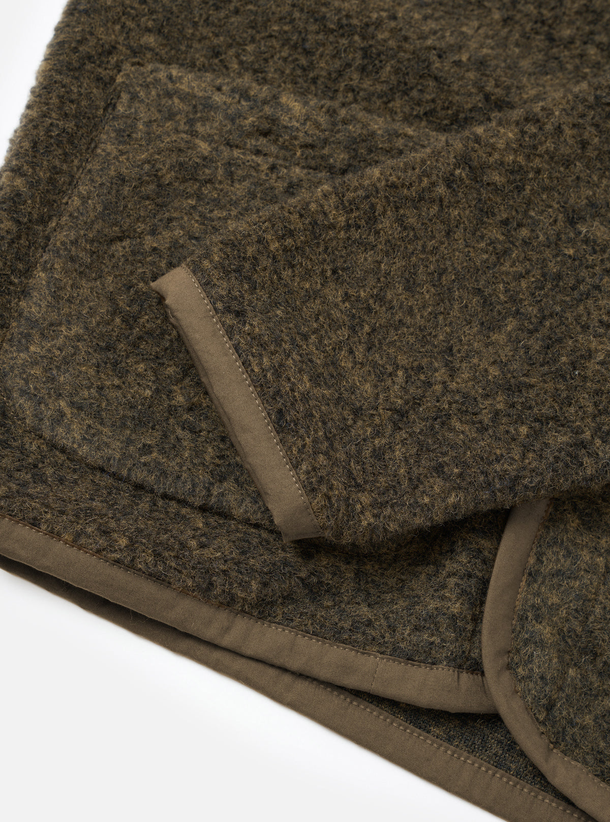 Close-up flat lay of a brown boucle style fleece Lancaster jacket on a white studio surface. The shot focuses on the folded hem and cuff, showing the jacket's thick, textured boucle style exterior and a contrasting tan woven binding trim with visible topstitching along the edges.