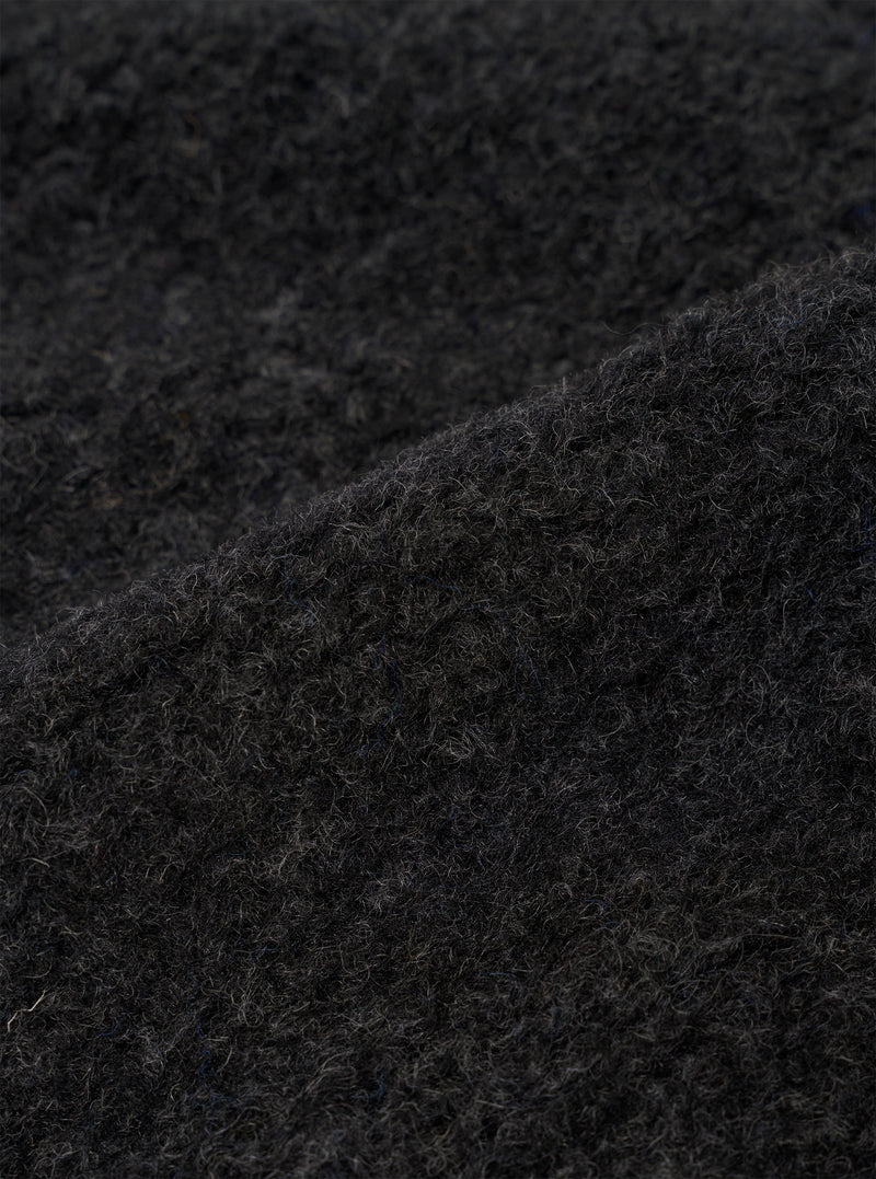 Close-up detail of the charcoal boucle style fleece fabric used on the Lancaster Jacket. The shot shows the dense, curly looped texture of the boucle style weave in dark charcoal grey, with individual fibres visible across the surface. A folded edge runs diagonally across the frame, highlighting the fabric's lofty, textured depth.