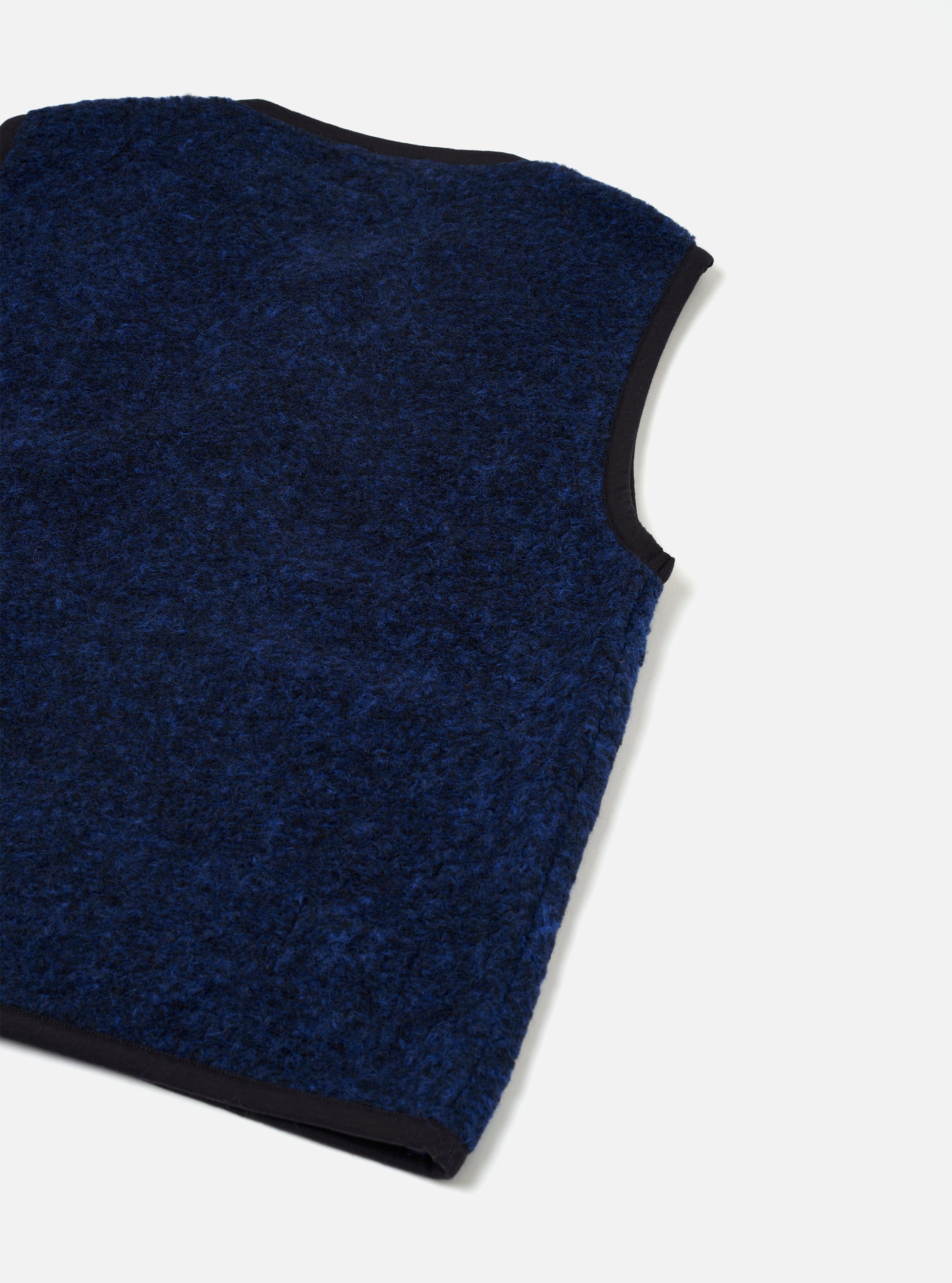 Close-up flat lay of a blue boucle style fleece gilet on a light grey seamless backdrop. The textured, looped boucle style fabric is visible across the body, with a navy binding trim running along the armhole and neckline edges. The hem and lower body are cropped into frame at an angle.