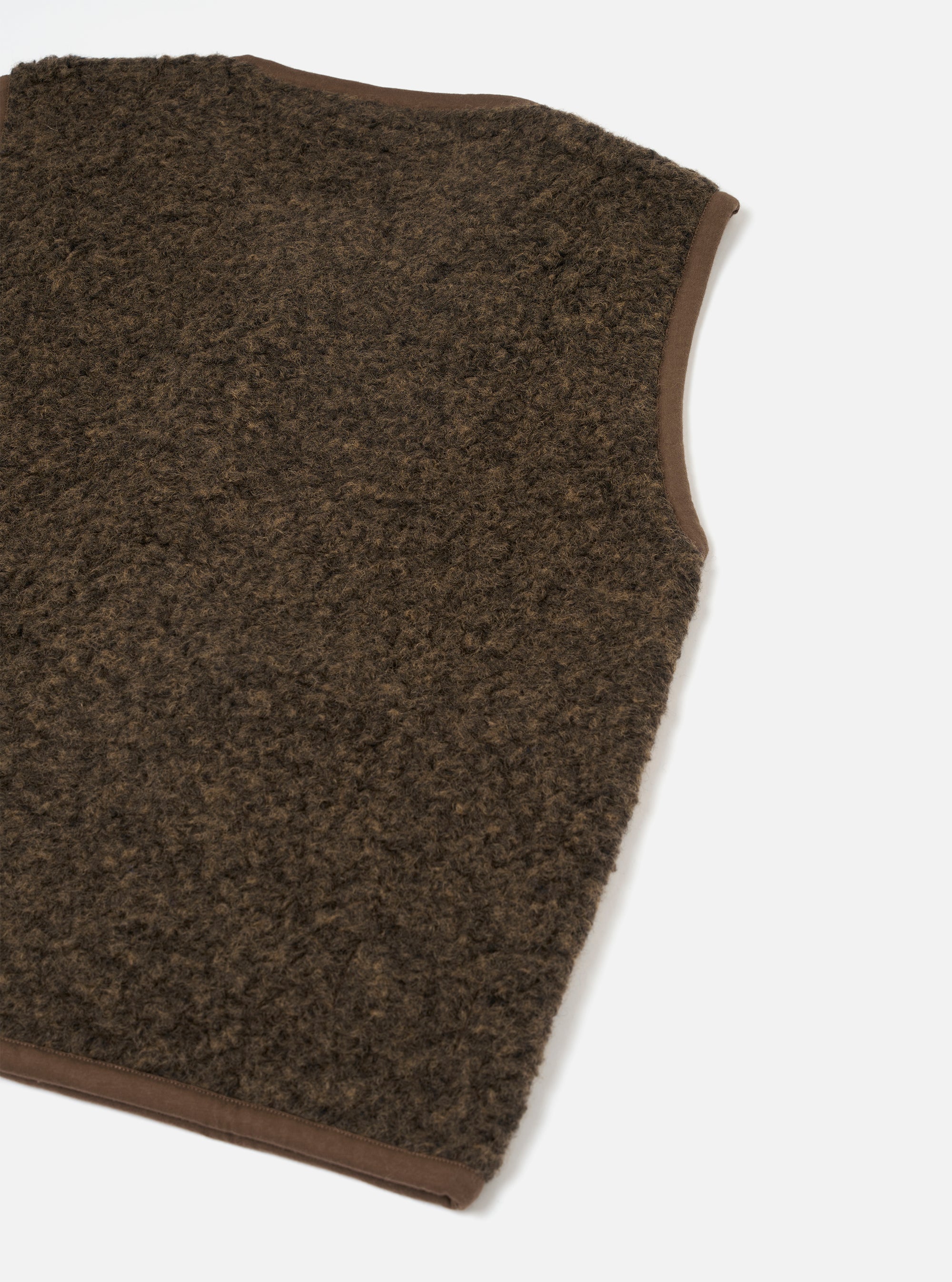 Close-up flat lay of a brown boucle style fleece gilet on a white studio backdrop. The dense, curly-textured fleece body is trimmed with smooth brown fabric binding along the armhole and hem edges. The shot is cropped to show the front panel, shoulder, and lower hem corner.