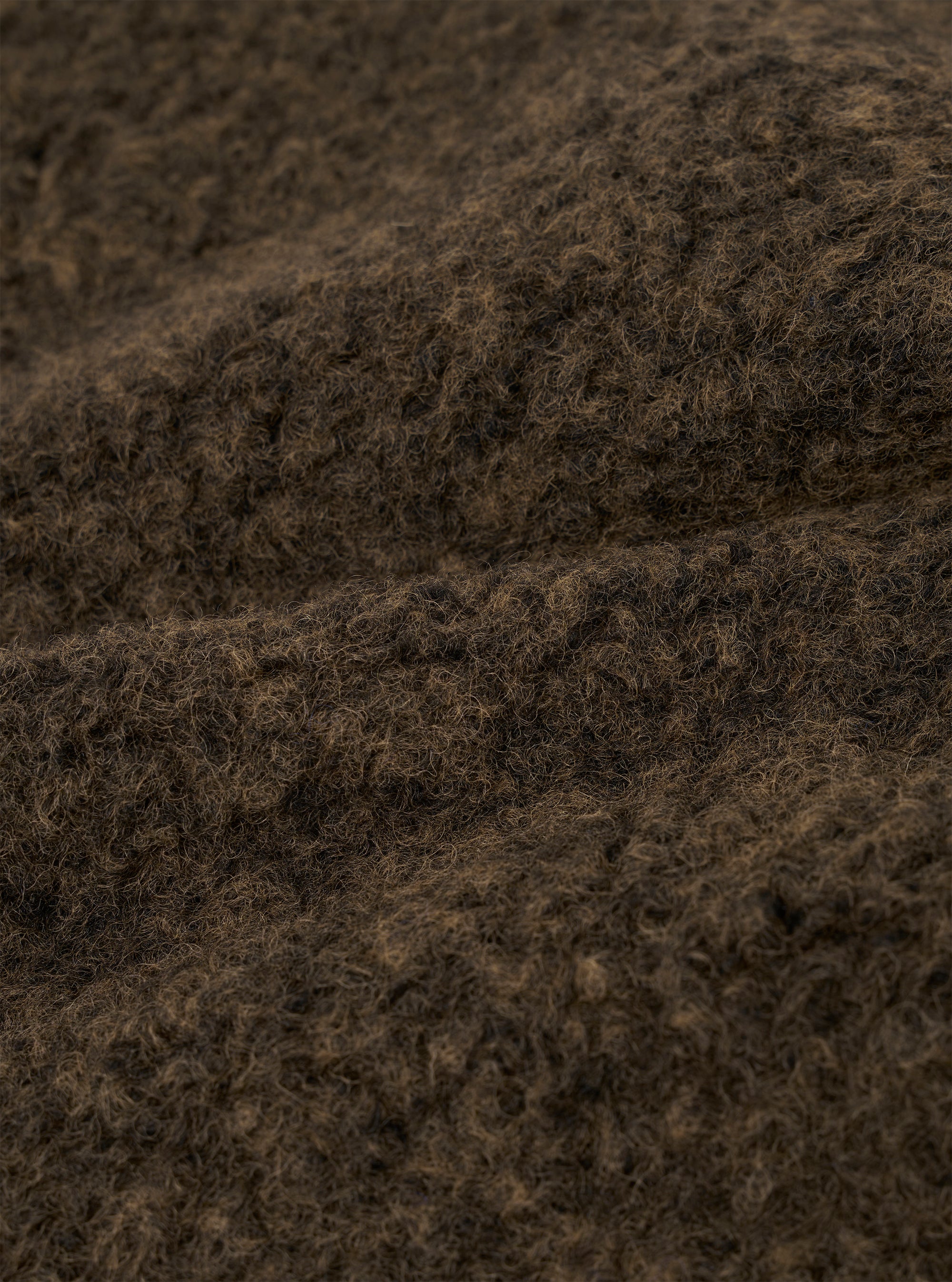 Close-up detail shot of the brown boucle style fleece fabric used in the Lancaster Gilet. The tightly looped, curly texture is clearly visible, with dark brown and warm tan tones running through the fibres. The image shows the fabric folded, highlighting its depth and soft, fluffy surface.