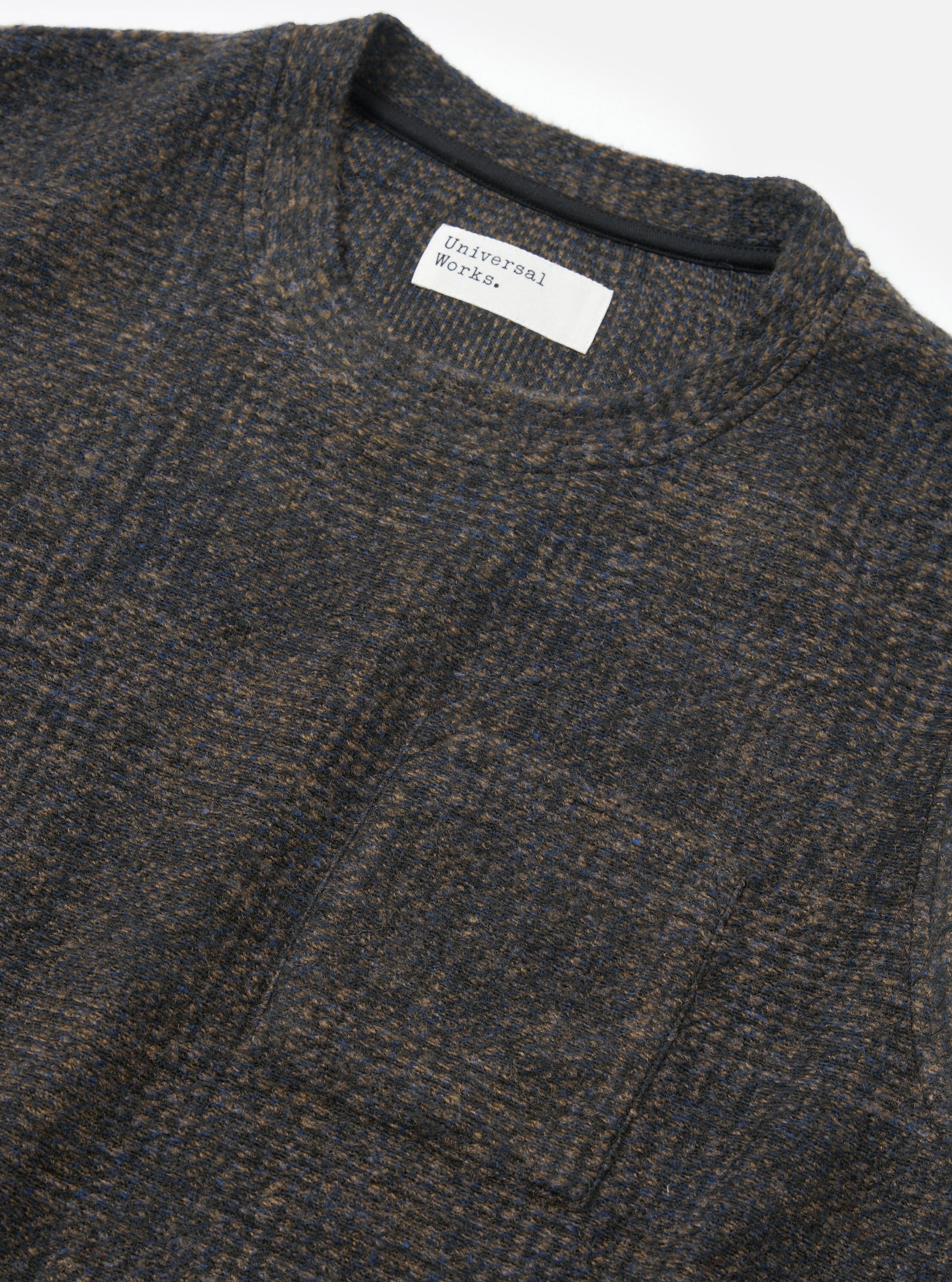 Close-up flat lay of a Universal Works navy Deca Light Knit Sleeveless Crew on a white studio surface. The marl fabric blends navy, brown and blue tones in a fine knit texture. A ribbed crew neckline is visible with a dark inner tape, and a woven Universal Works label sits at the front neck.