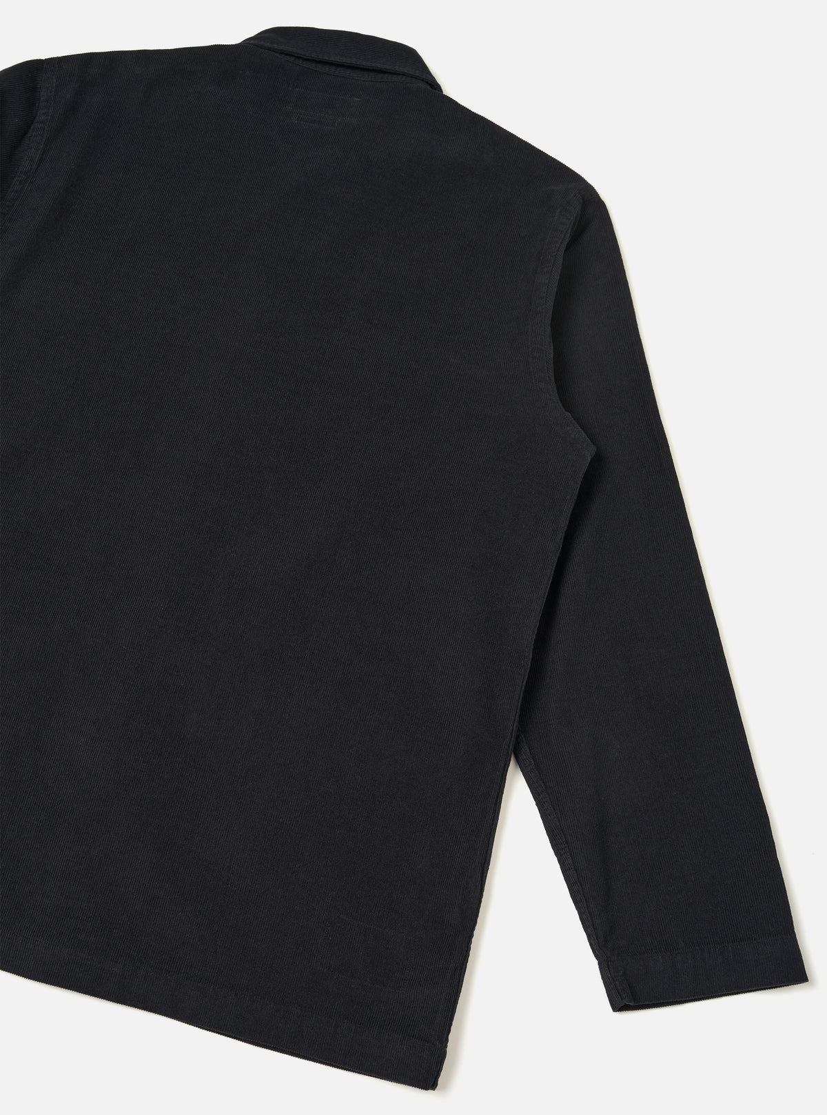 Rear studio view of the Black Fine Cord Bakers Overshirt against a clean white backdrop. The photo highlights the flat back yoke, straight hemline, and fine-ribbed black corduroy fabric construction.