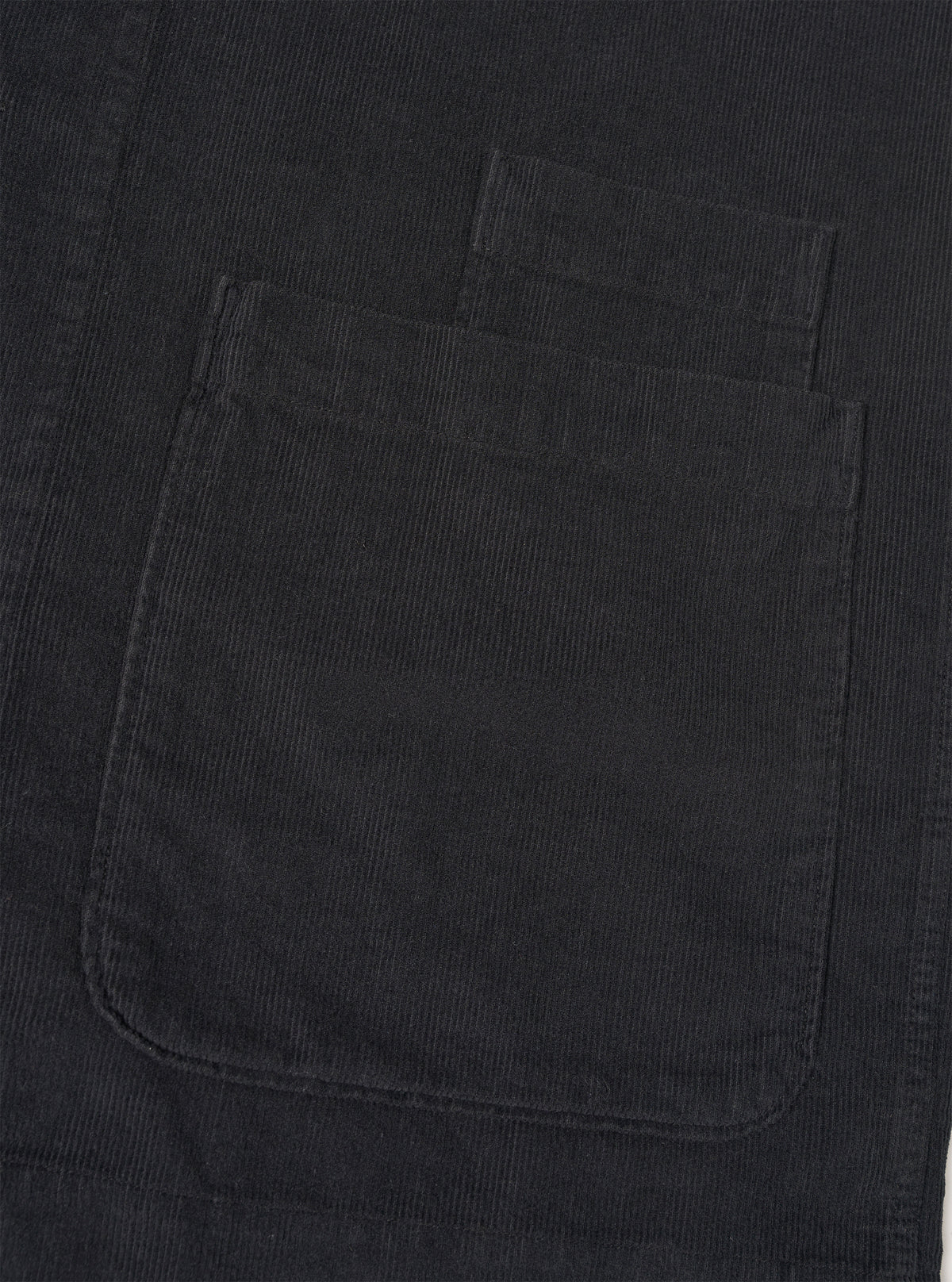 Detailed close-up shot of the Black Fine Cord Bakers Overshirt on a white background, highlighting the chest patch pocket, fine corduroy texture, button detail, and neat edge stitching on the black garment.