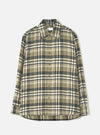 A male model wearing the Universal Works Olive Jazz Check Standard Shirt, standing against a plain off-white background, displaying the classic button-down style, olive checked pattern, and crisp collar.