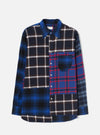 Blue patchwork flannel shirt laid flat on a light grey backdrop. Four contrasting check panels are cut and sewn together: navy and white on the left body and sleeve, blue and red tartan on the right body, and grey and blue check on the right sleeve. Point collar, button-down placket with silver buttons, single chest patch pocket, and a curved hem.