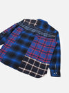 Blue patchwork flannel shirt laid flat on a light grey seamless backdrop, shot from above at an angle showing the back. Multiple check panels in blue, black, red and white are pieced together across the body and sleeves. A patch pocket sits at centre back. The cuff features two silver buttons.