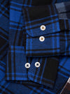 Close-up flat lay of a blue and black patchwork check flannel shirt. Two cuffs are visible, each fastened with white four-hole buttons. The upper cuff has one button, the lower cuff has two. Brushed twill fabric shows bold cobalt and black tartan checks across contrasting patchwork panels.
