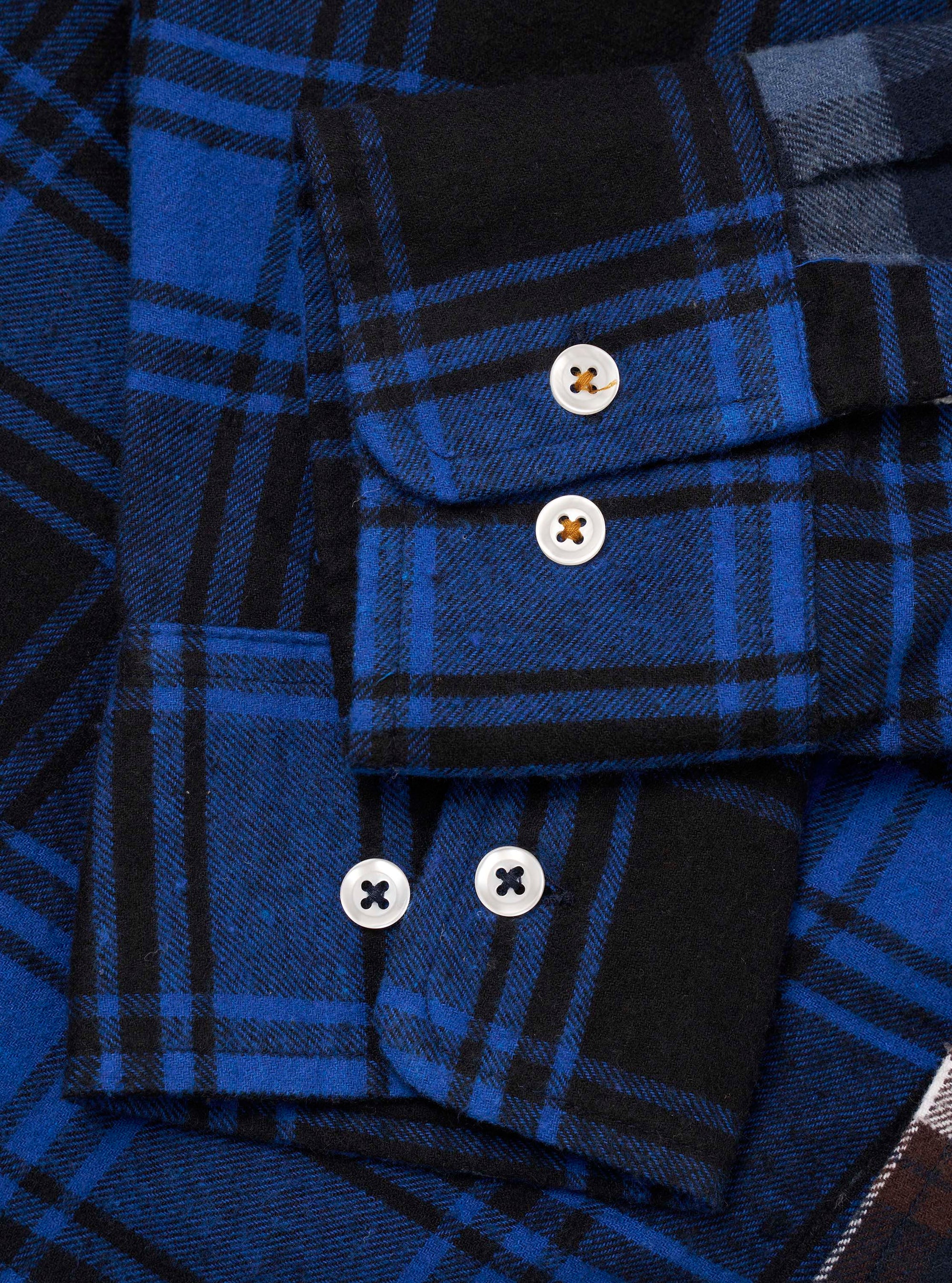 Close-up flat lay of a blue and black patchwork check flannel shirt. Two cuffs are visible, each fastened with white four-hole buttons. The upper cuff has one button, the lower cuff has two. Brushed twill fabric shows bold cobalt and black tartan checks across contrasting patchwork panels.
