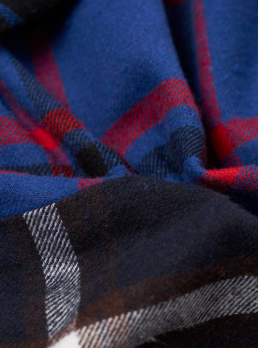 Close-up detail shot of a blue patchwork checks shirt, showing two contrasting check panels. One panel features a bold blue ground with red and black tartan lines; the other shows a dark charcoal ground with white and grey checks. The woven wool-effect fabric has a soft, brushed texture visible throughout.